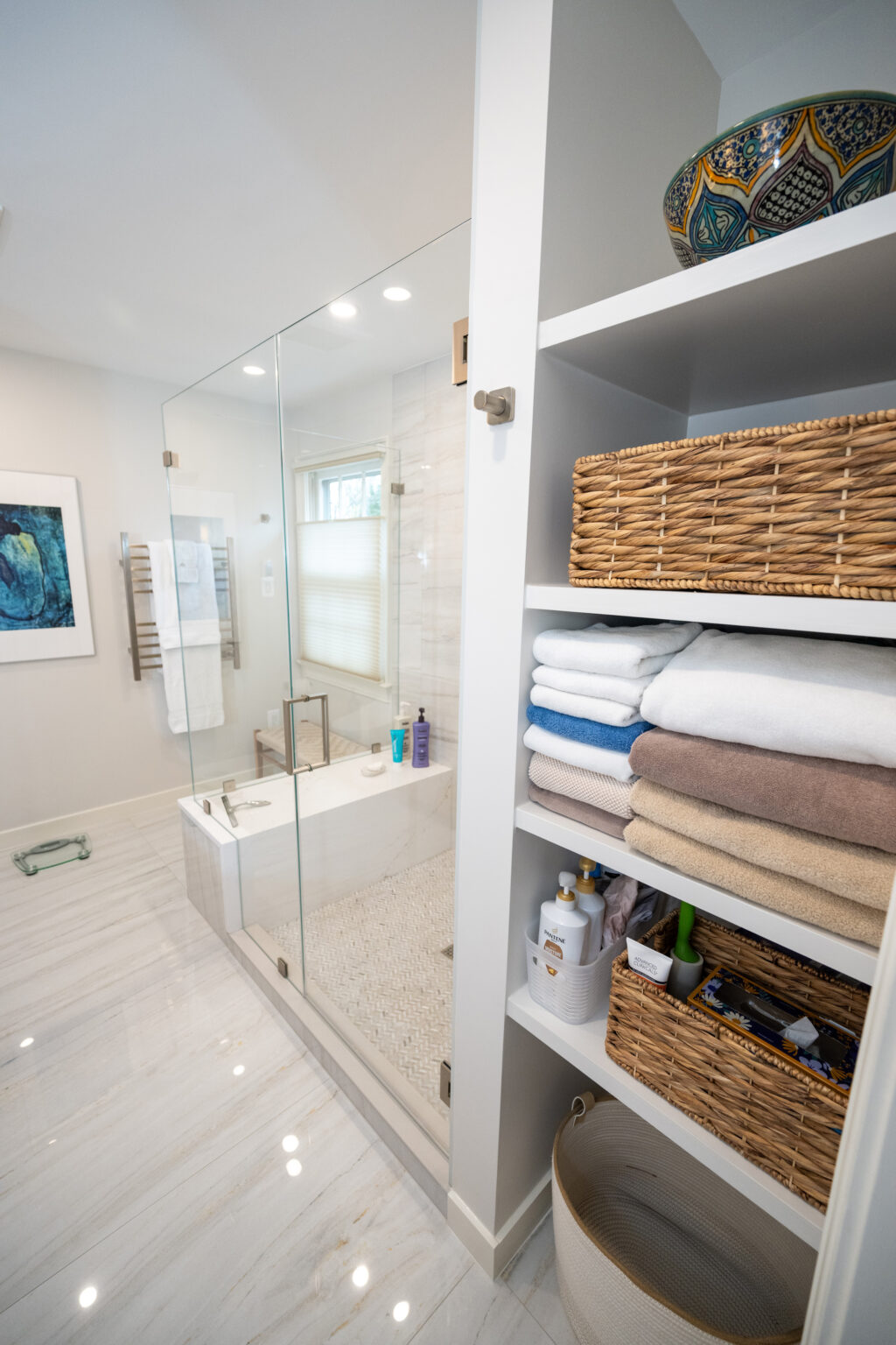 Bathroom featuring a glass-enclosed shower with built-in bench, tiled walls, and floor. Nearby, there are open shelves with neatly stacked towels, baskets, and toiletries. A piece of art hangs on the wall.
