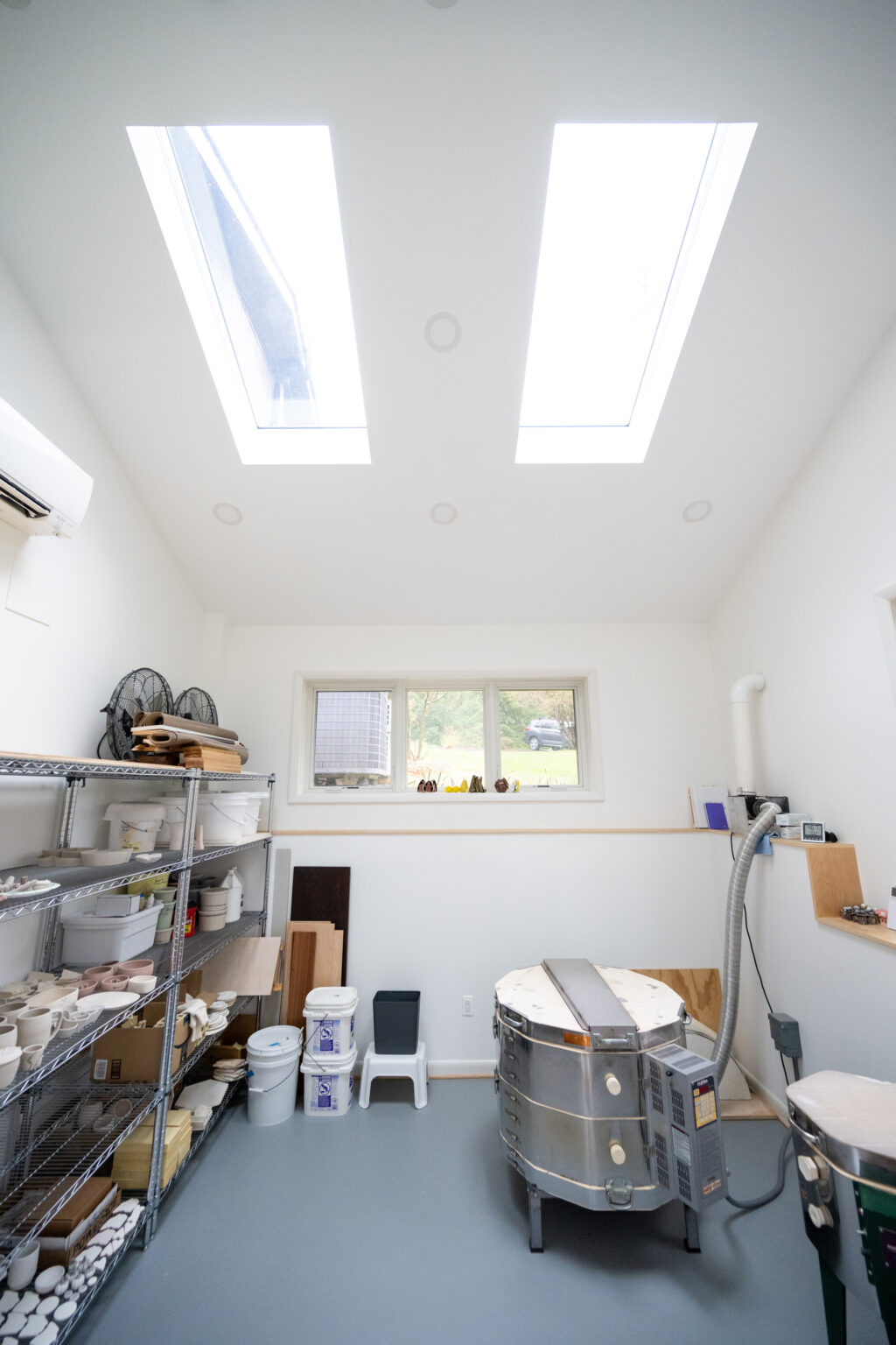 A bright pottery studio with natural light from skylights, shelves with supplies on the left, a windowed wall at the back, and a pottery kiln on the right.