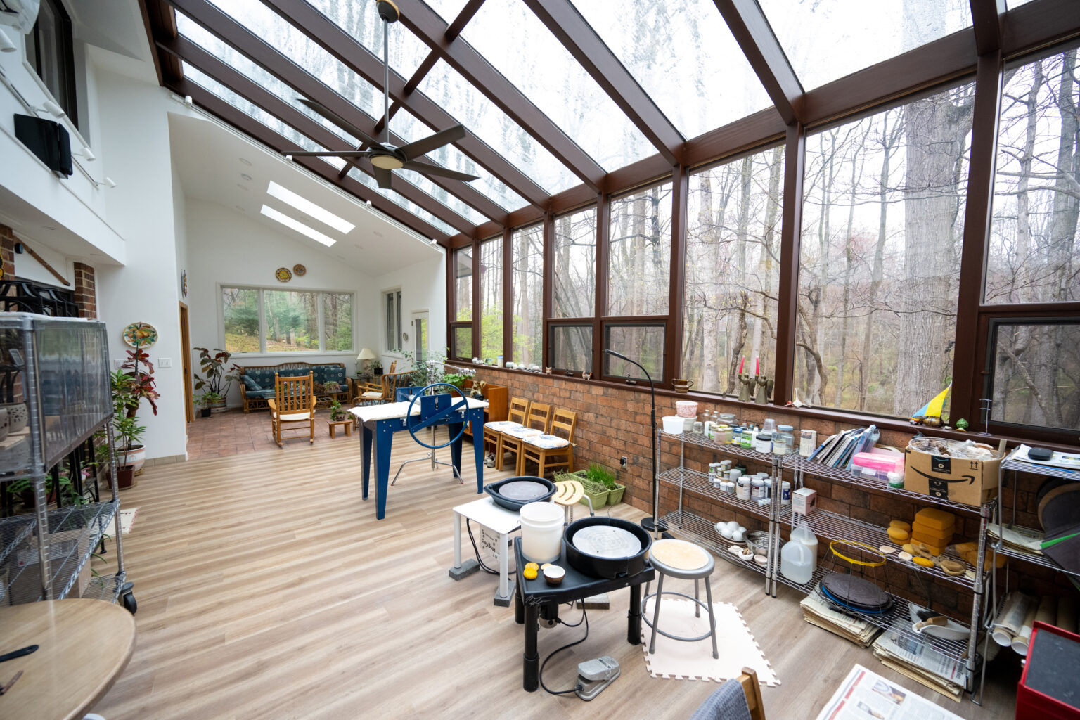 A sunlit room with large glass windows, housing pottery equipment including wheels, shelves with tools and supplies, and various pieces of furniture. Trees and forest seen outside the windows.