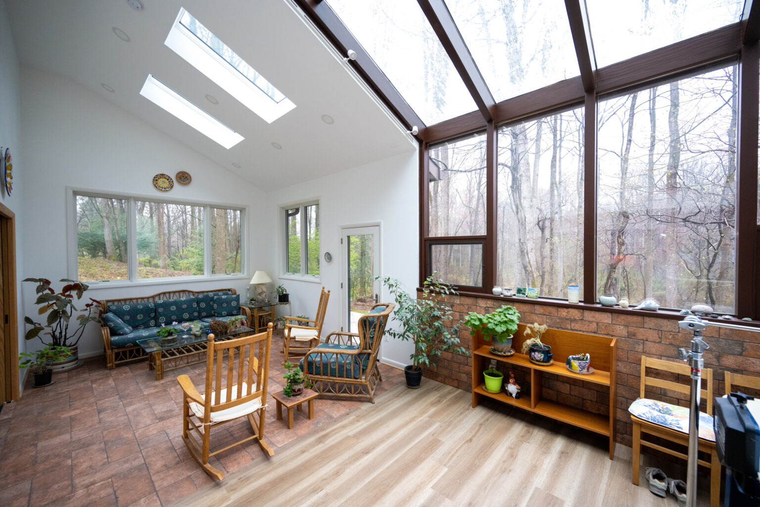 A sunlit conservatory with large windows, furnishing includes wooden chairs, sofas, plants, and shelves. The floor is a mix of wood and tile, and the outside view shows bare trees.