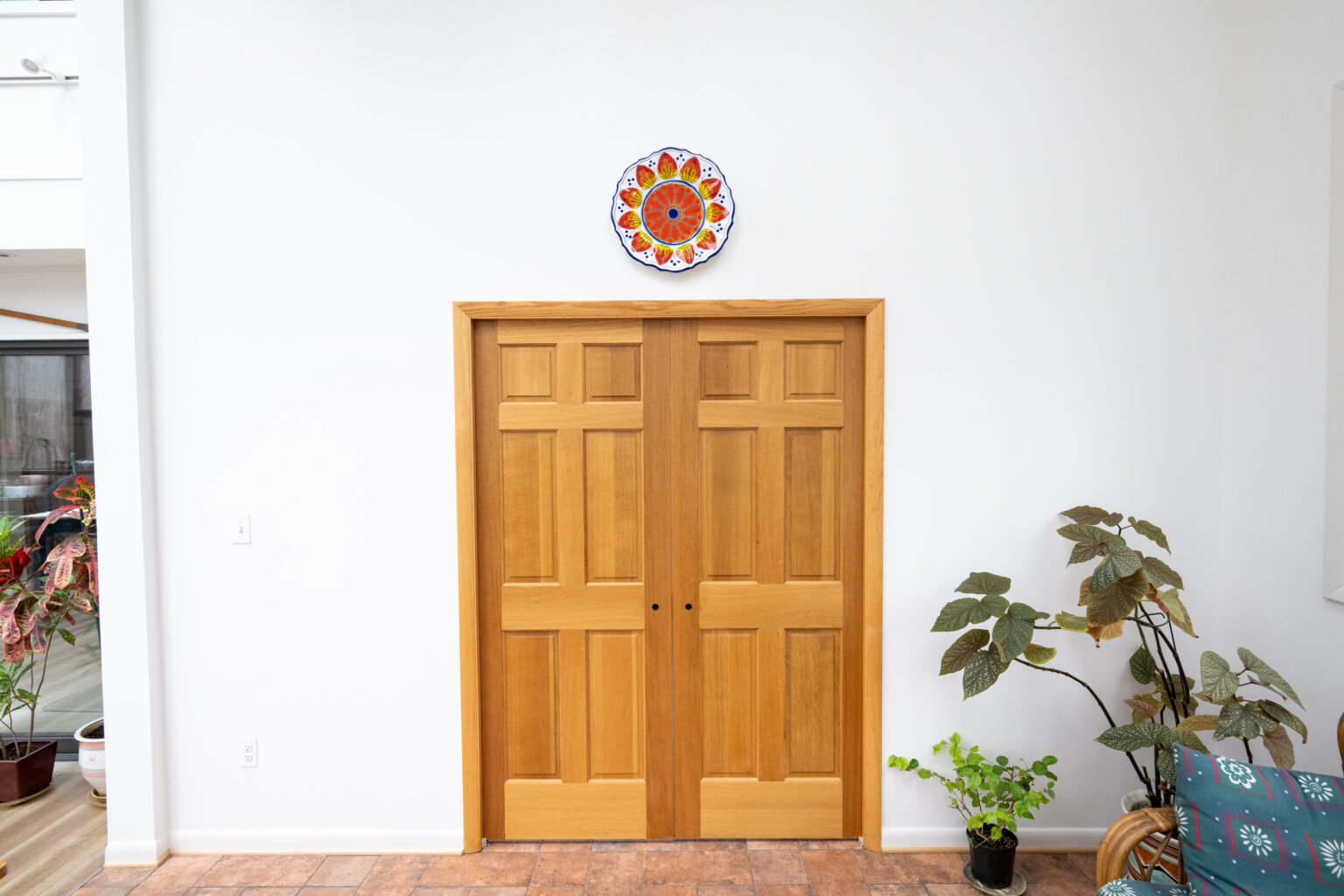 A pair of wooden double doors in a white room, with a colorful circular wall decor above. Green plants are positioned on either side of the doors.