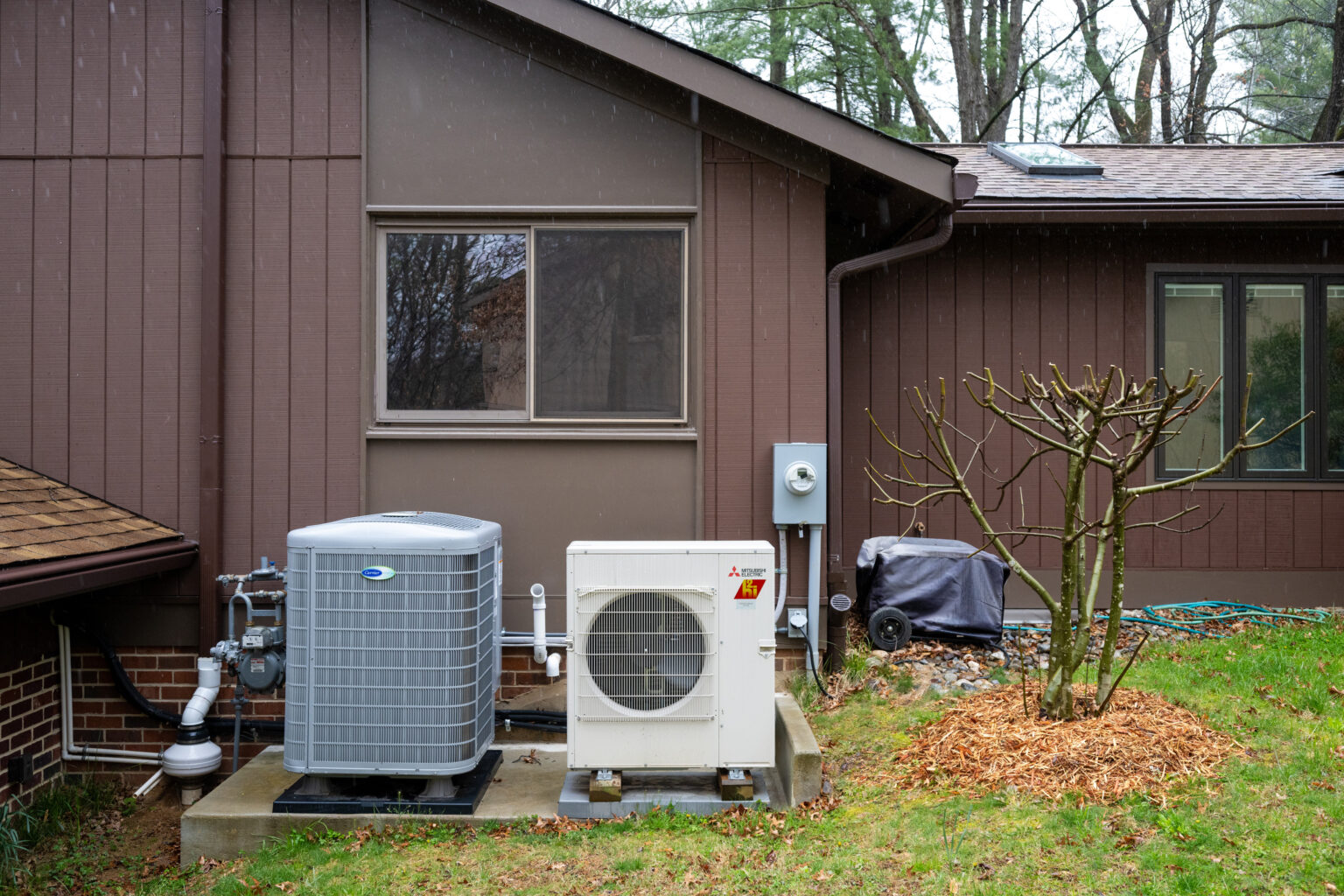 Outdoor air conditioning and heating units placed on a concrete pad next to a brown house, with a bare tree and covered grill in the yard.