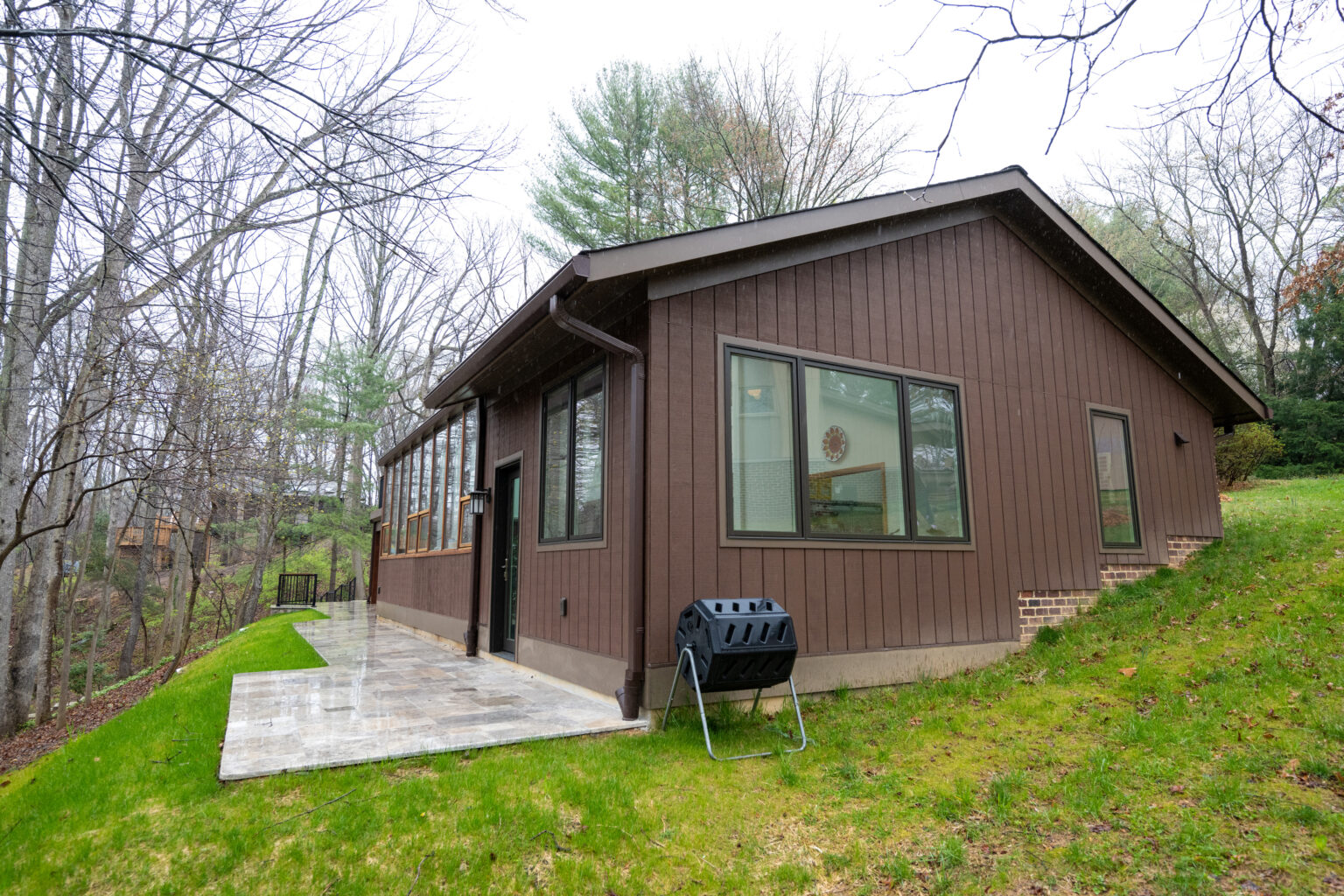 A brown, single-story house with large windows and a concrete pathway surrounded by grass and trees on a cloudy day. A black grill is placed on the lawn near the house.