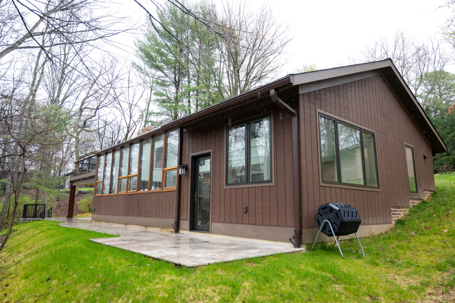 A brown wooden house with large windows sits on a grass lawn surrounded by trees. A patio with a grill is visible on the side. It appears to be a single-story home.
