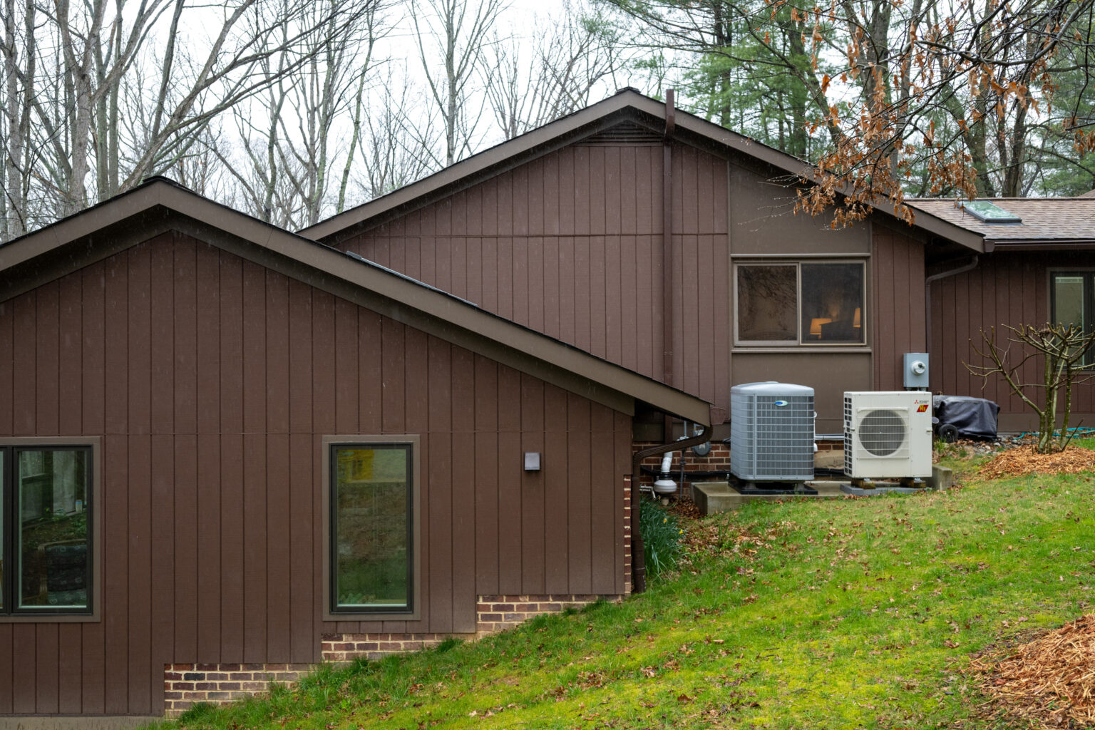 Two brown residential buildings with large windows. The yard has green grass, leafy trees in the background, and HVAC units on the ground level.