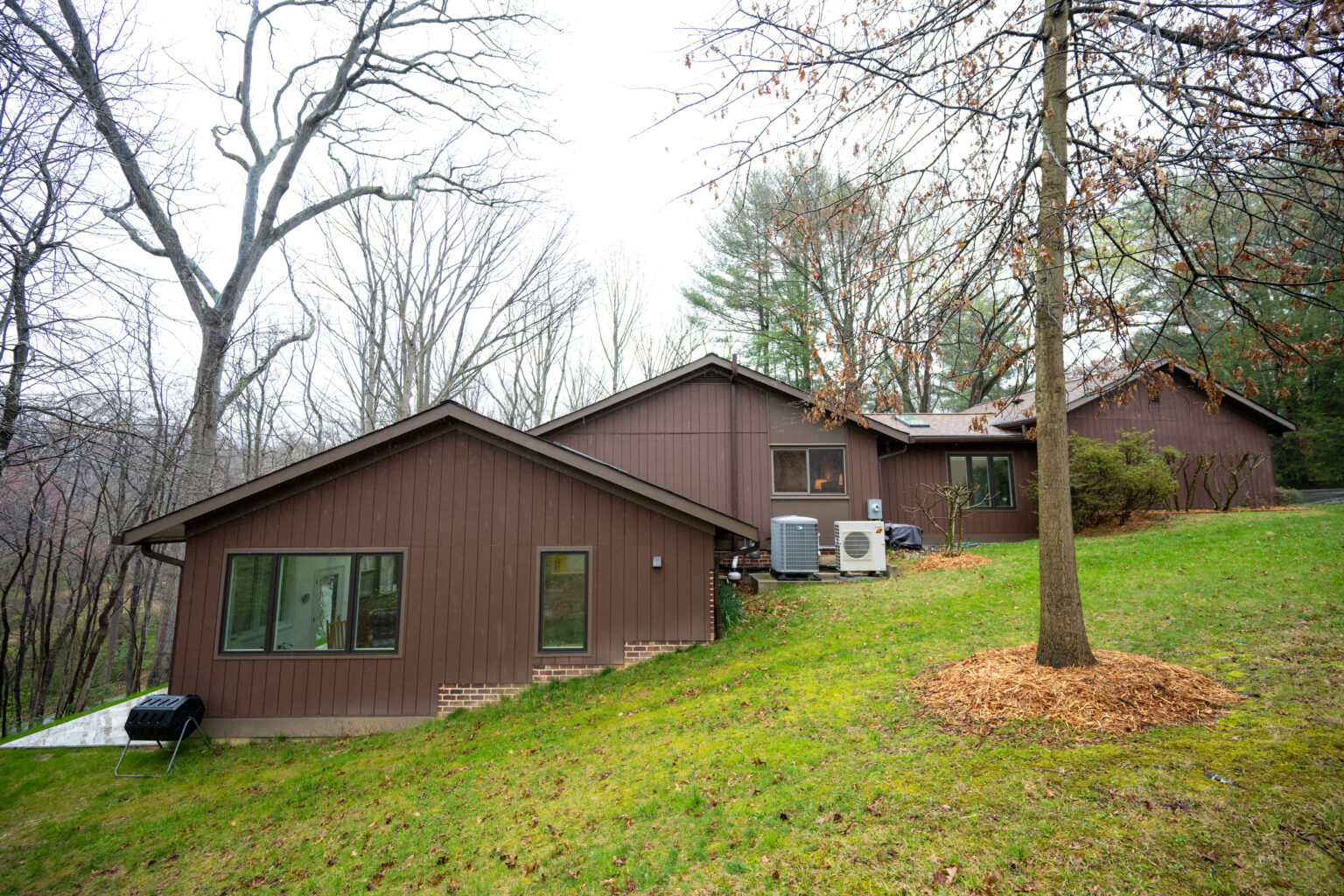A brown, multi-sectioned house sits on a grassy slope surrounded by leafless trees, with two air conditioning units and a grill visible in the backyard.