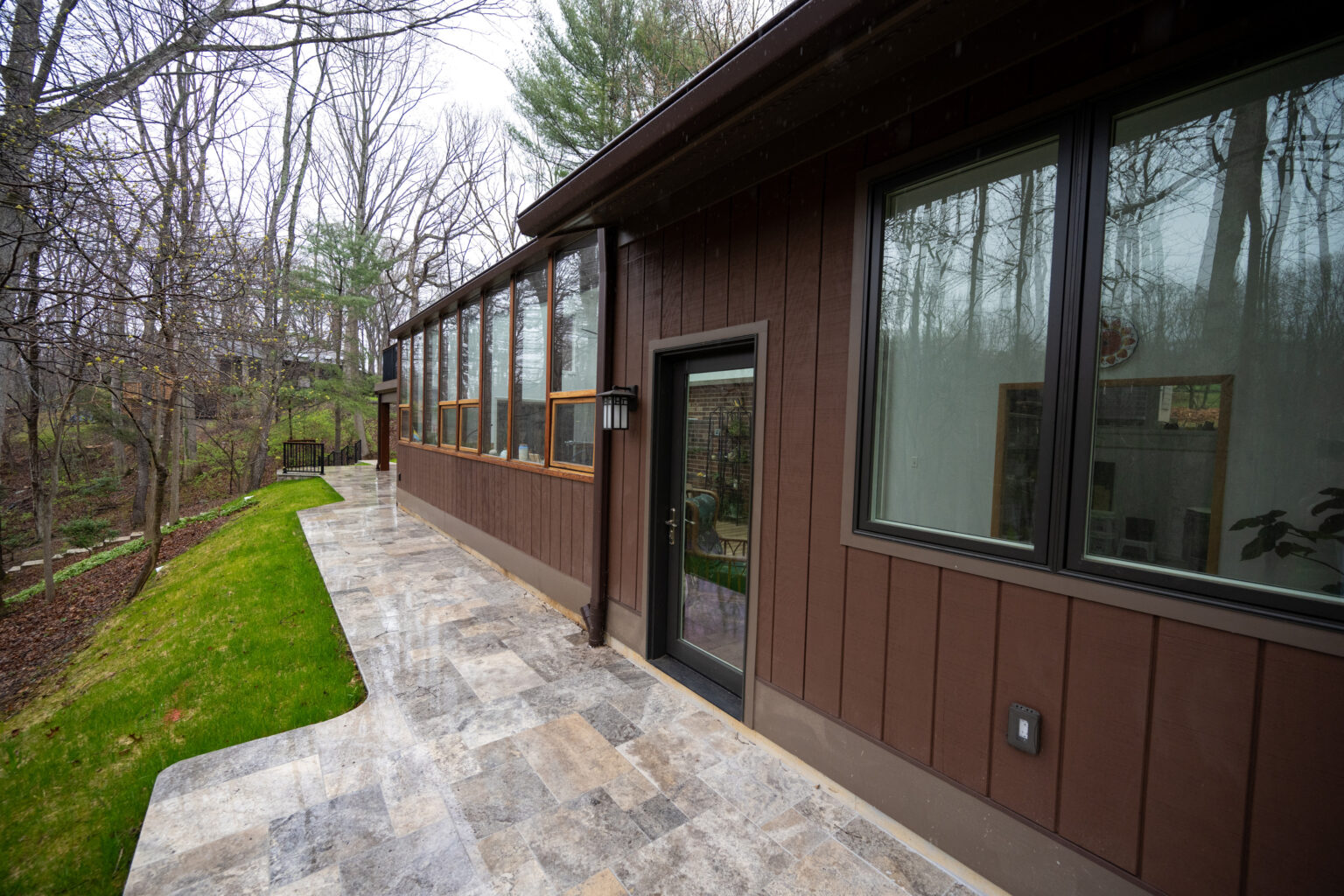 A modern house with large windows and a stone walkway is located in a wooded area during early spring. The ground near the walkway is covered with grass and fallen leaves.