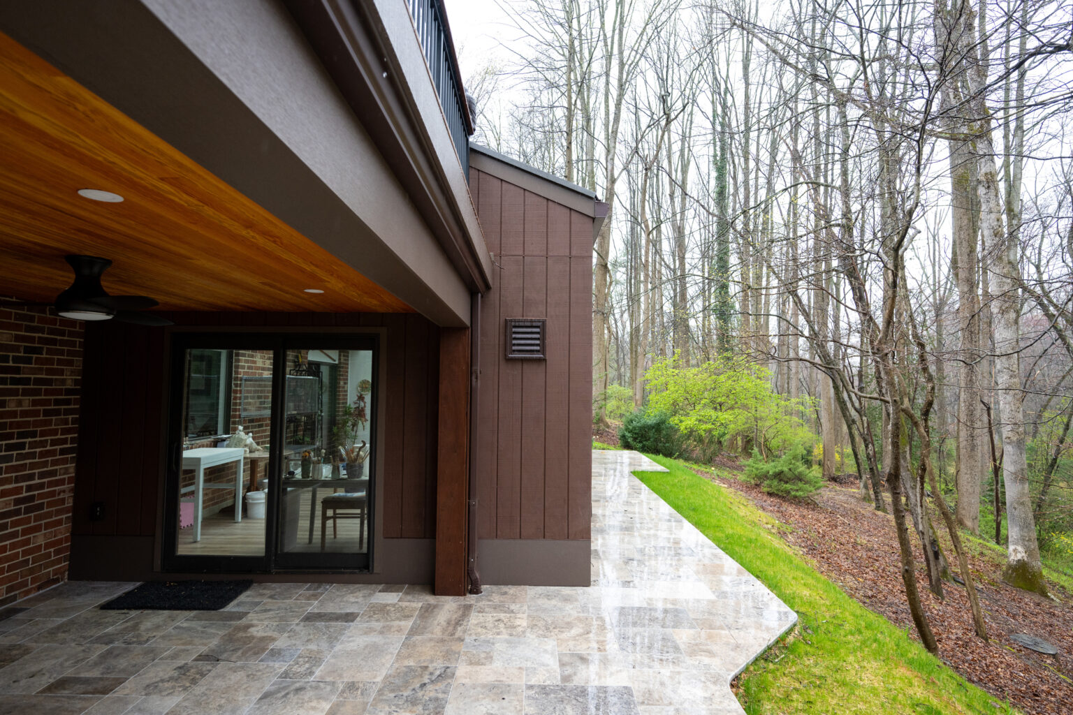 Rear view of a house with a covered patio, sliding glass doors, and a pathway leading into a wooded area with bare trees. A ceiling fan is mounted on the patio ceiling.