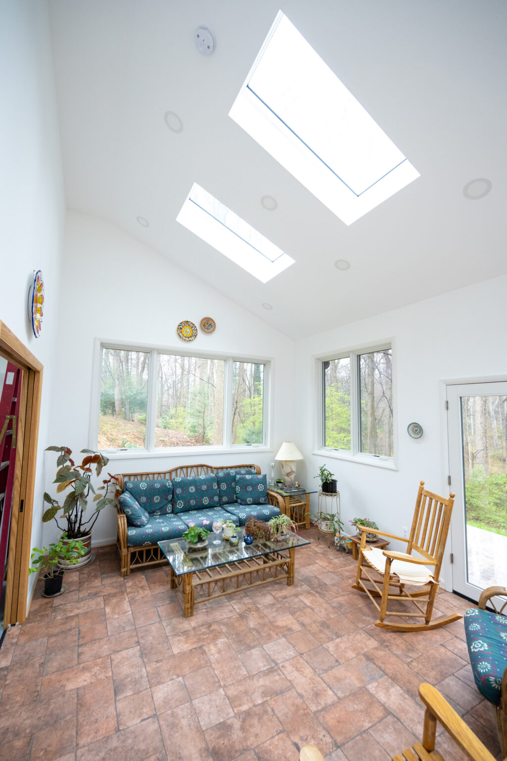 A bright, white-walled sunroom with skylights, tiled floor, two rocking chairs, a patterned sofa, various plants, and decorative wall plates. Large windows offer a view of the wooded outside area.