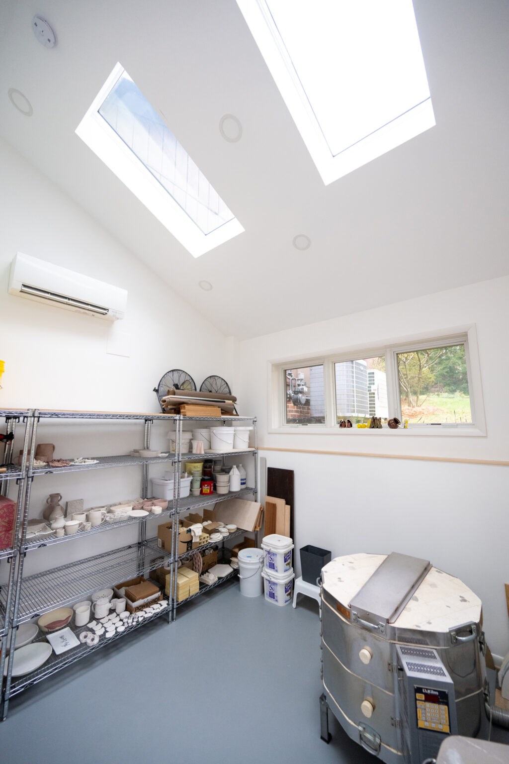 A pottery studio with two skylights, shelves filled with pottery supplies, and a ceramic kiln. A window offers an outdoor view.