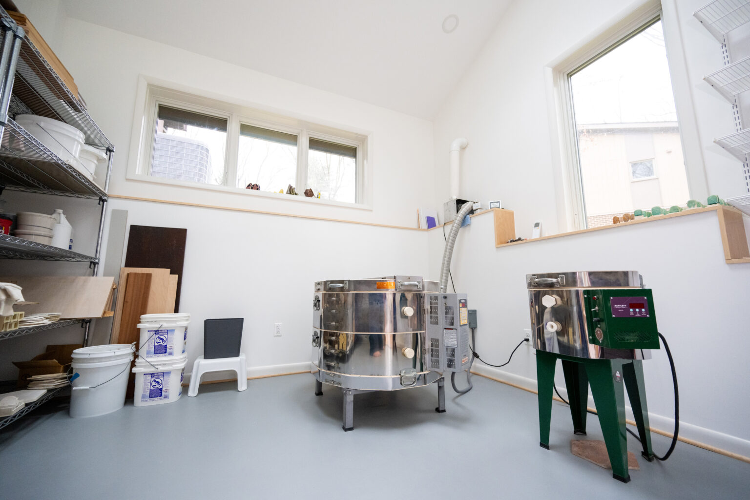 A well-lit pottery studio with two kilns, various shelves holding pottery supplies, and a few small, finished pots visible on a window ledge.