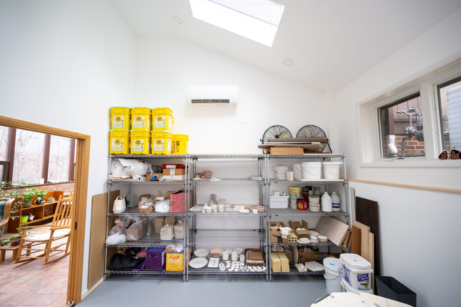 A well-organized storage room with metal shelves holding various supplies, including yellow buckets, white containers, and pottery items. A skylight and a window provide natural light.