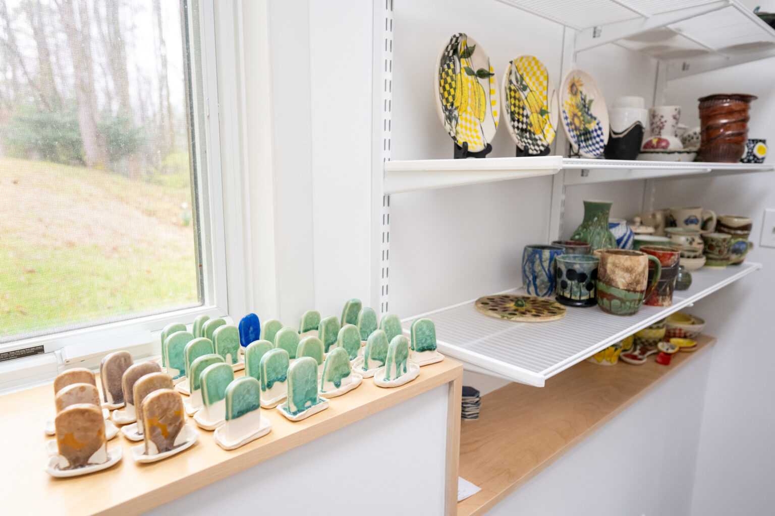 Shelves displaying various ceramic items, including colorful plates, mugs, vases, and a collection of small cactus-shaped figurines, against a white wall with a window to the left.