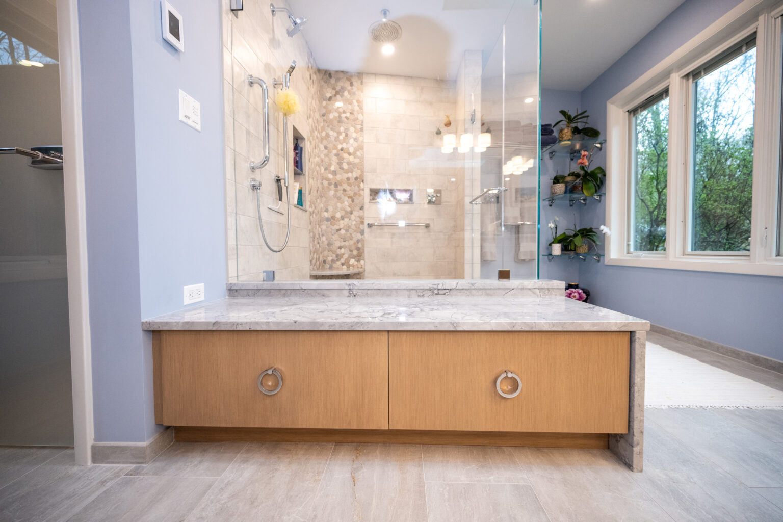 Image by Lesnick Photo Modern bathroom with a large vanity featuring wooden cabinets and a marble countertop. The glass-enclosed shower area has beige tiles and multiple showerheads. A window allows natural light into the room.