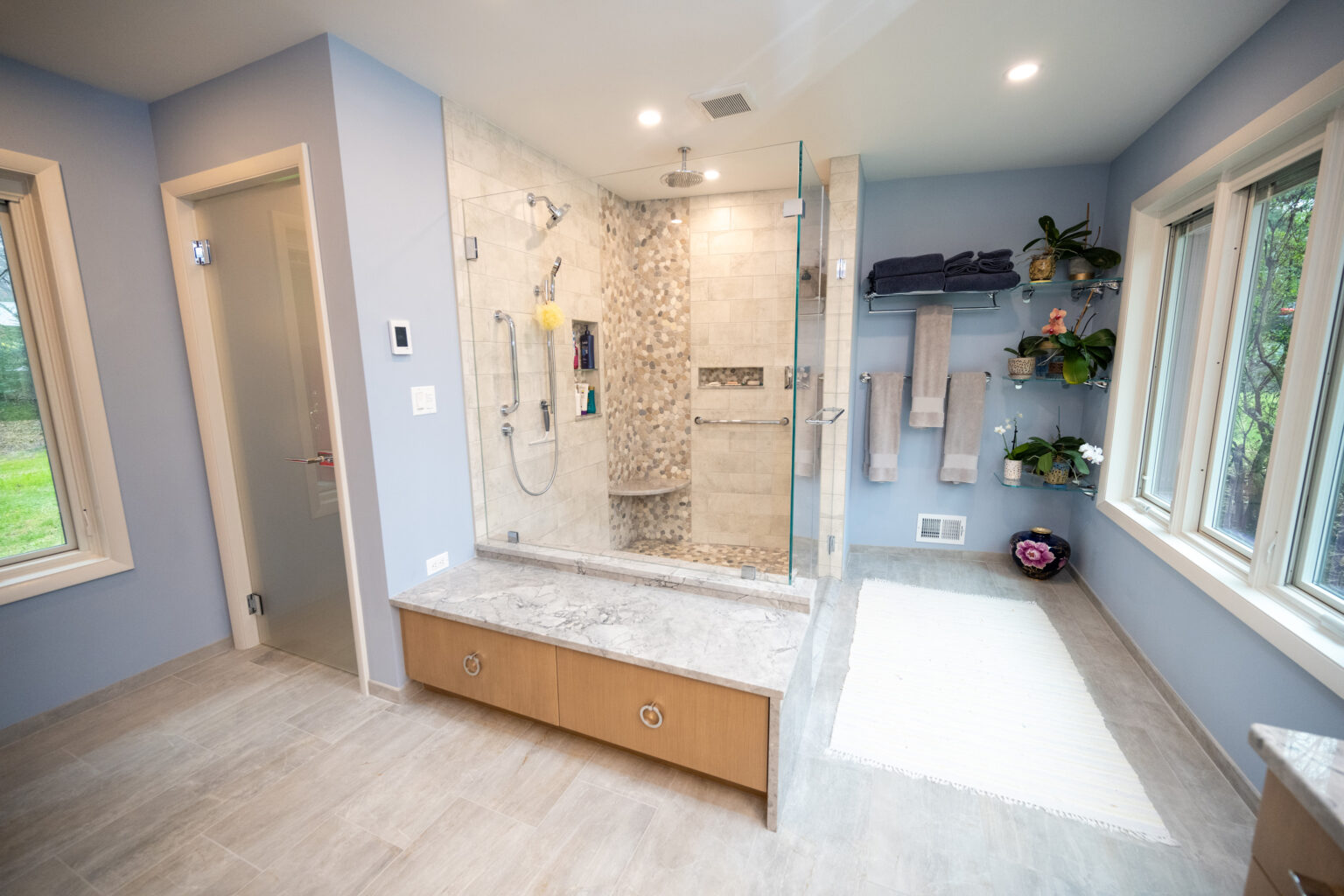 Image by Lesnick Photo A modern bathroom with a glass-enclosed shower, beige tiles, built-in seating, and a shelf with towels and plants. The room features large windows, light-colored flooring, and a wall-mounted control panel.