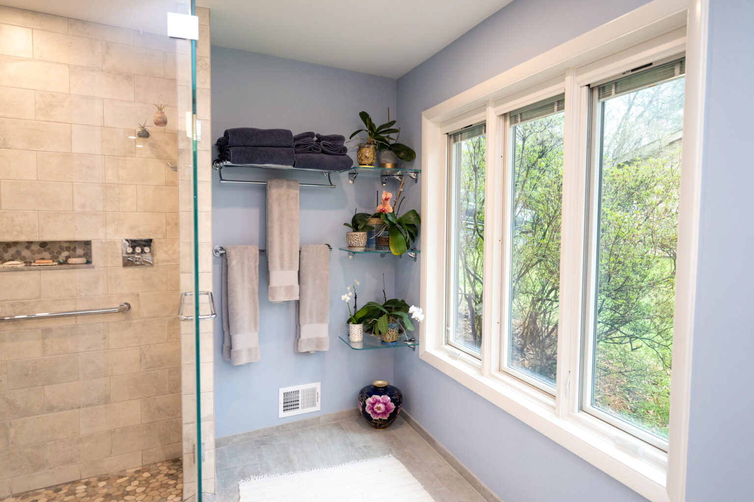 Image by Lesnick Photo A bathroom with light blue walls, a glass shower, shelves with towels and plants, a window with a view of greenery, and a small rug on the floor.