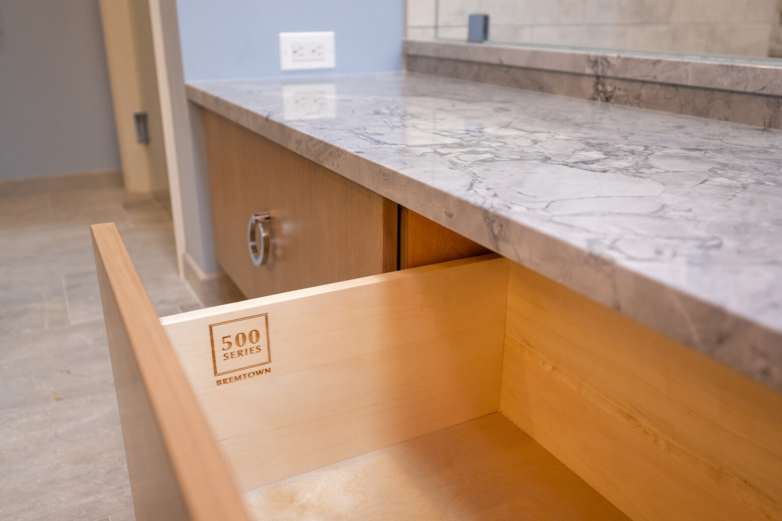 Image by Lesnick Photo Open wooden drawer in a bathroom vanity showcasing "500 Series" Bremtown logo on the side, with a marble countertop, wall outlet, and mirror in the background.