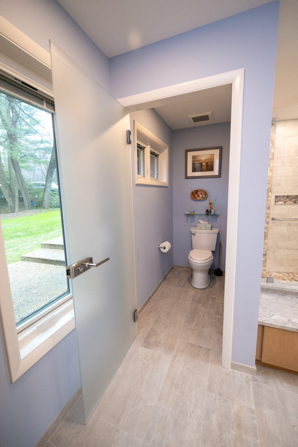 Image by Lesnick Photo A bathroom with blue walls, a frosted glass door, a window, a toilet, toilet paper holder, and a countertop with a sink. A picture is hung above the toilet, and the floor is tiled.