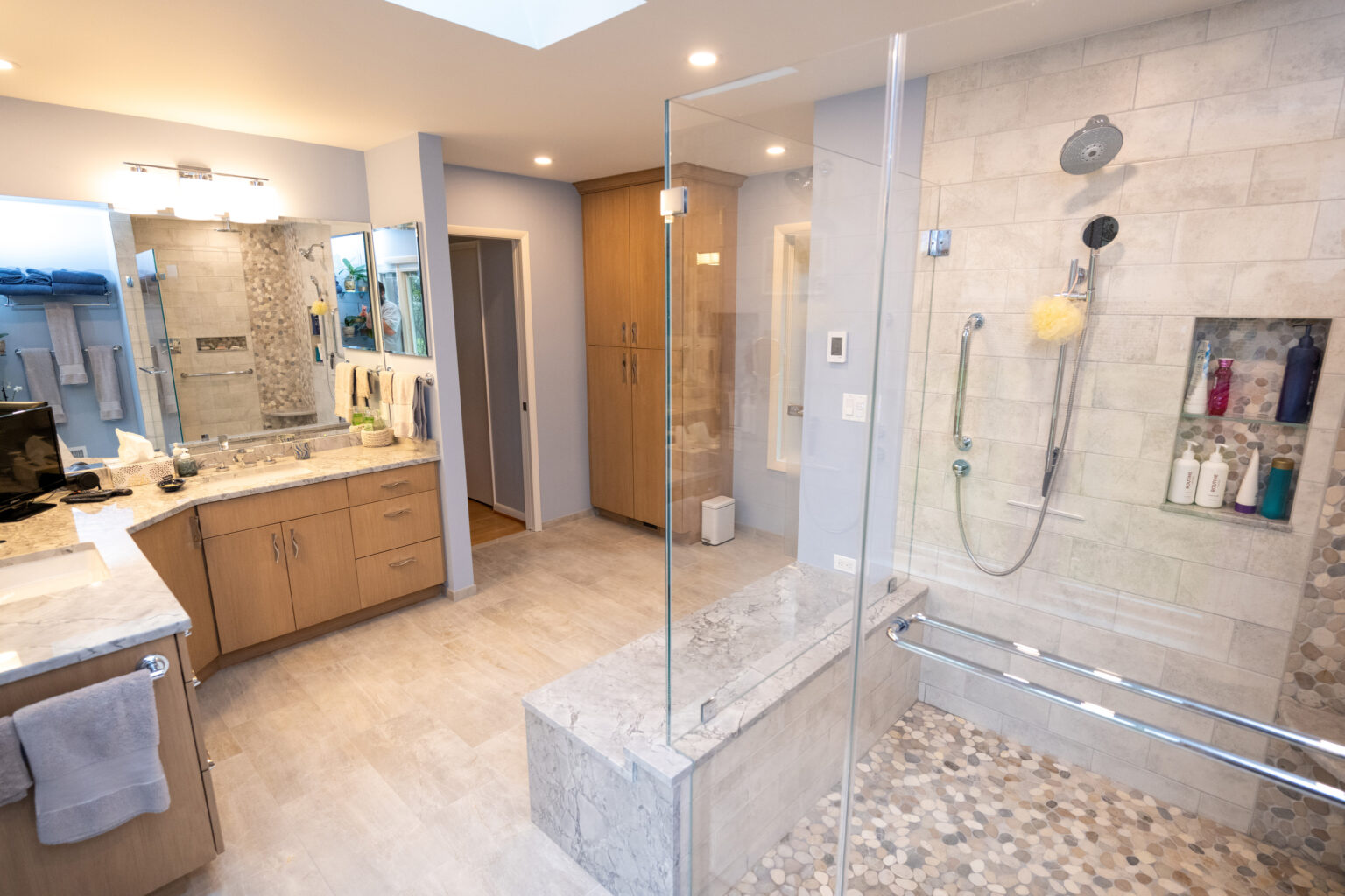 Image by Lesnick Photo A modern bathroom featuring a glass-enclosed shower with a bench, light wood cabinets, a large mirror, and a double sink vanity.