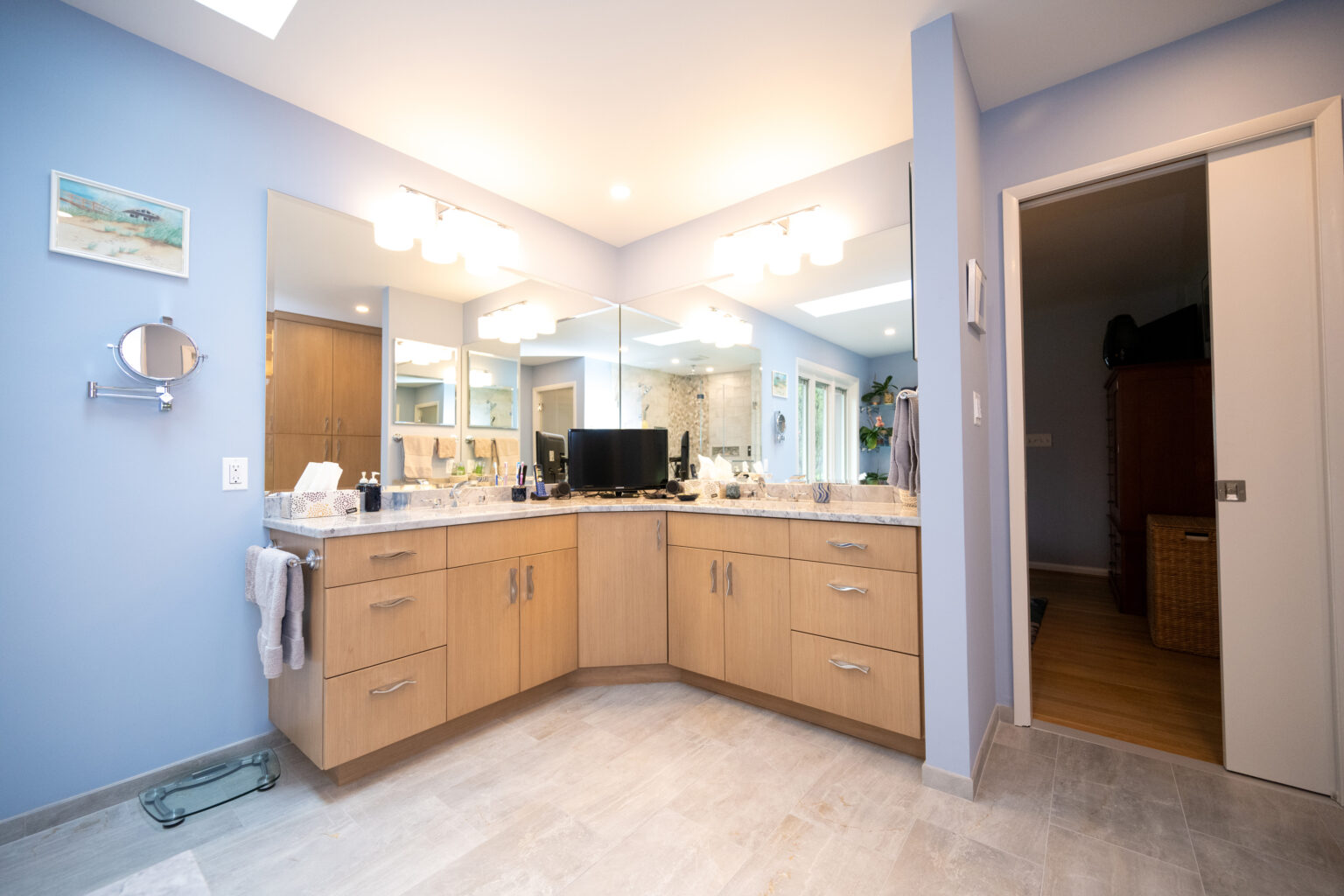 Image by Lesnick Photo A modern bathroom with blue walls features a wooden vanity with two sinks and a large mirror. Overhead lighting illuminates the space, and an adjacent room is visible through an open door.