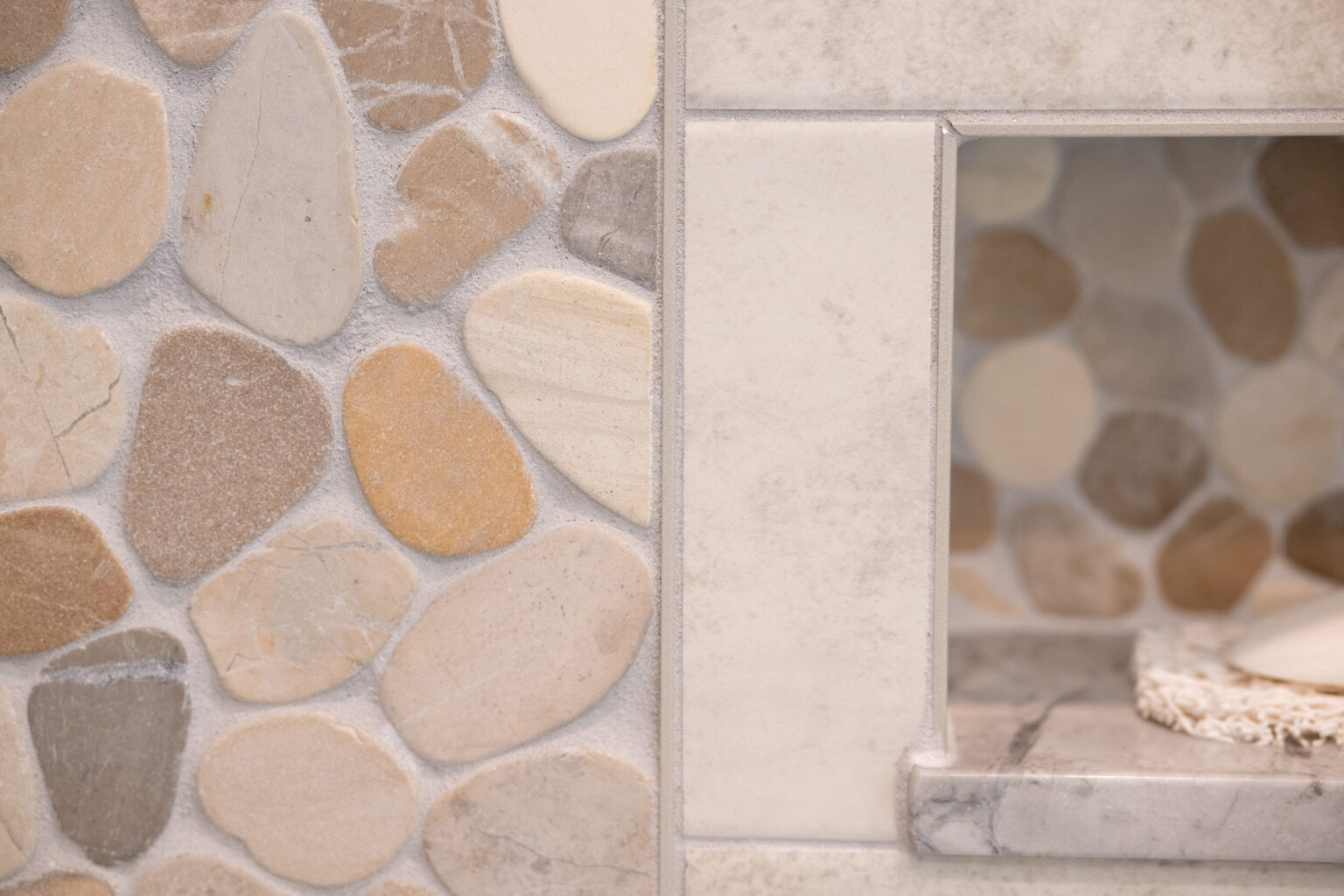 Image by Lesnick Photo Close-up of a wall featuring a pebble stone mosaic pattern on the left and a shelf with a similar stone design on the right. The stones are various shades of beige, light brown, and grey.