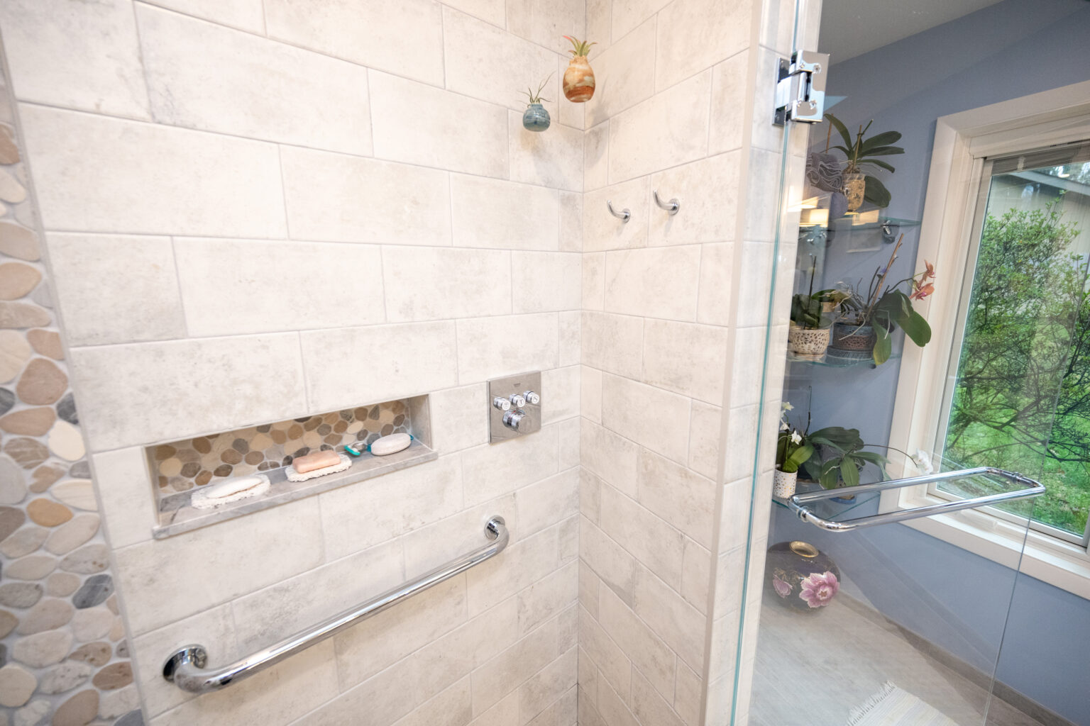 Image by Lesnick Photo A modern shower with beige tile walls, a built-in niche holding soap, a stainless steel grab bar, two hooks, and a glass door. The outside view shows a greenery through a window.