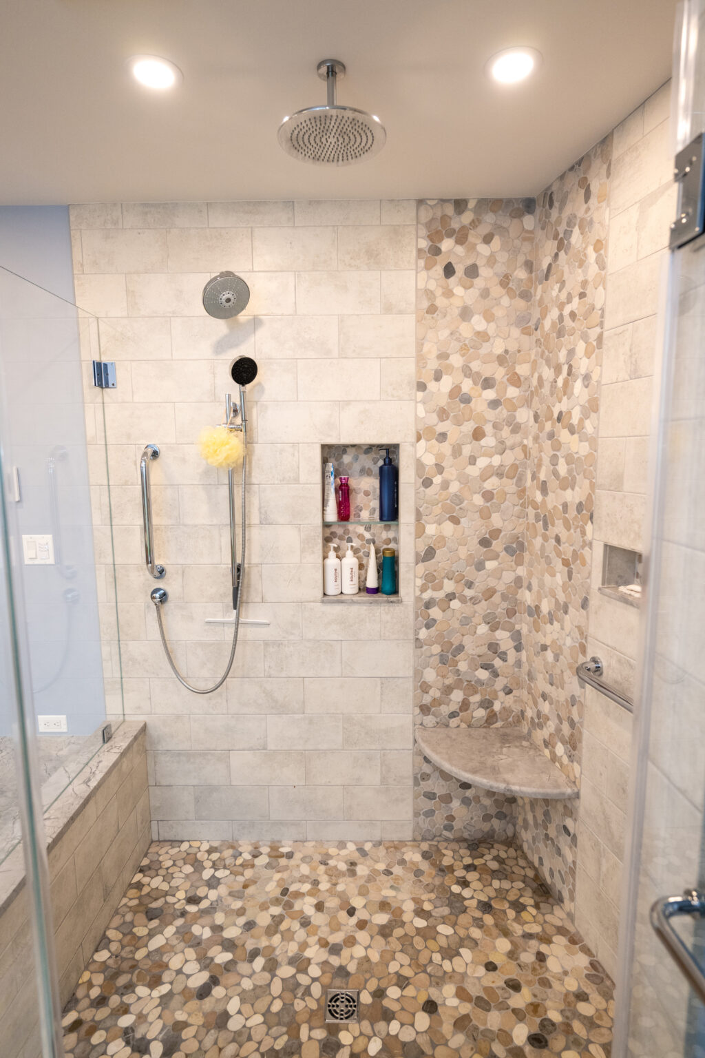 Image by Lesnick Photo Modern bathroom shower with beige and grey pebble tile floor and accent wall, tiled bench, niche with toiletries, handheld showerhead, overhead rain showerhead, and grab bars.