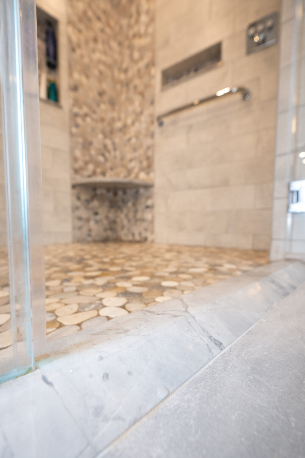 Image by Lesnick Photo Close-up view of a modern shower interior with pebble tile flooring, a built-in shelf with toiletries, and a wall-mounted grab bar.