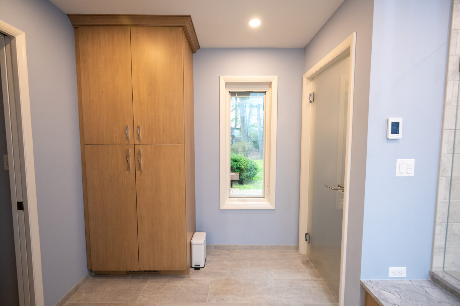 Image by Lesnick Photo A bathroom with light blue walls, a wooden cabinet, a small window, a white waste bin, a gray floor, and a closed glass door.