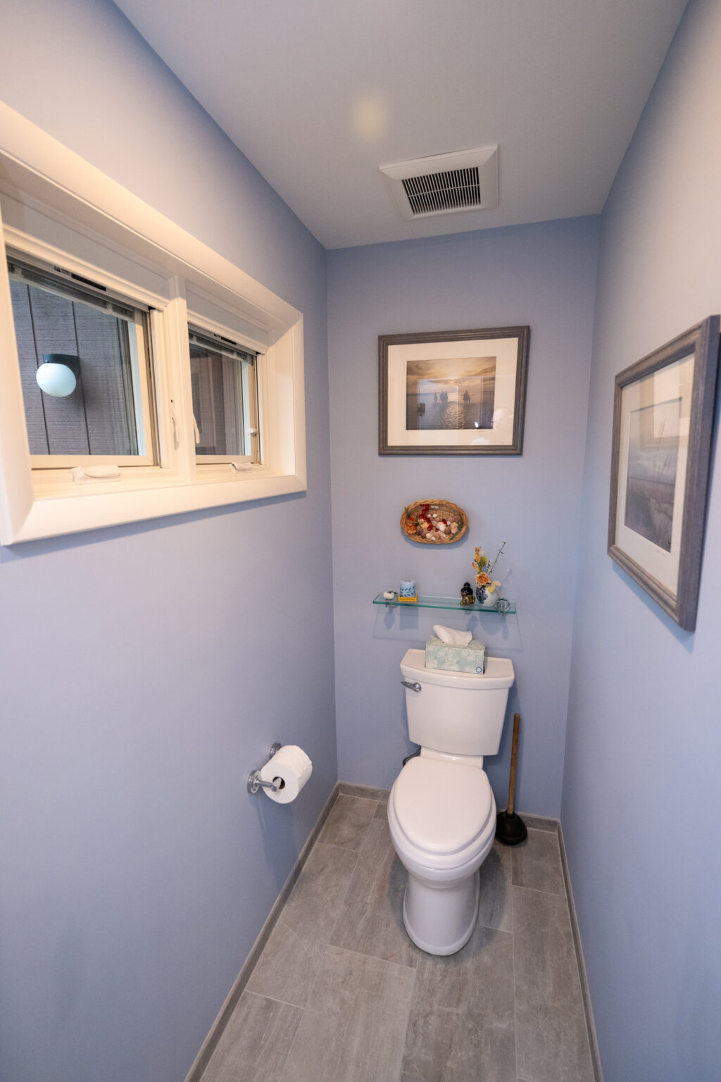 Image by Lesnick Photo A narrow bathroom with blue walls features a toilet, toilet paper holder, small shelf with tissue box, window, and framed artwork.