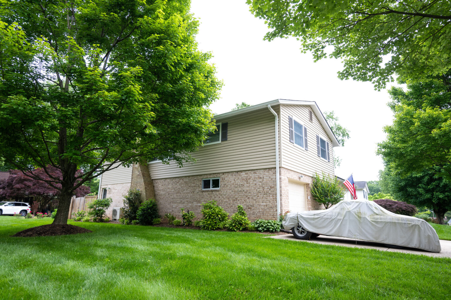 A house with beige siding and a brick lower section is surrounded by a neatly maintained lawn and trees. A car covered with a white tarp is parked in the driveway. An American flag is displayed near the house.