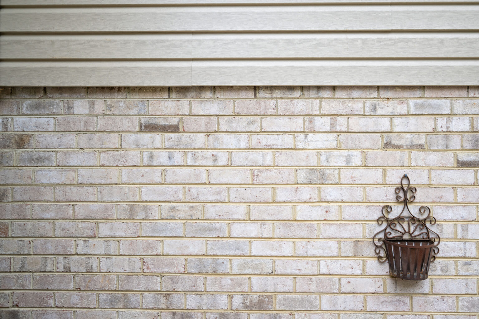 A decorative wall-mounted metal planter hangs on a brick wall beneath beige horizontal siding.
