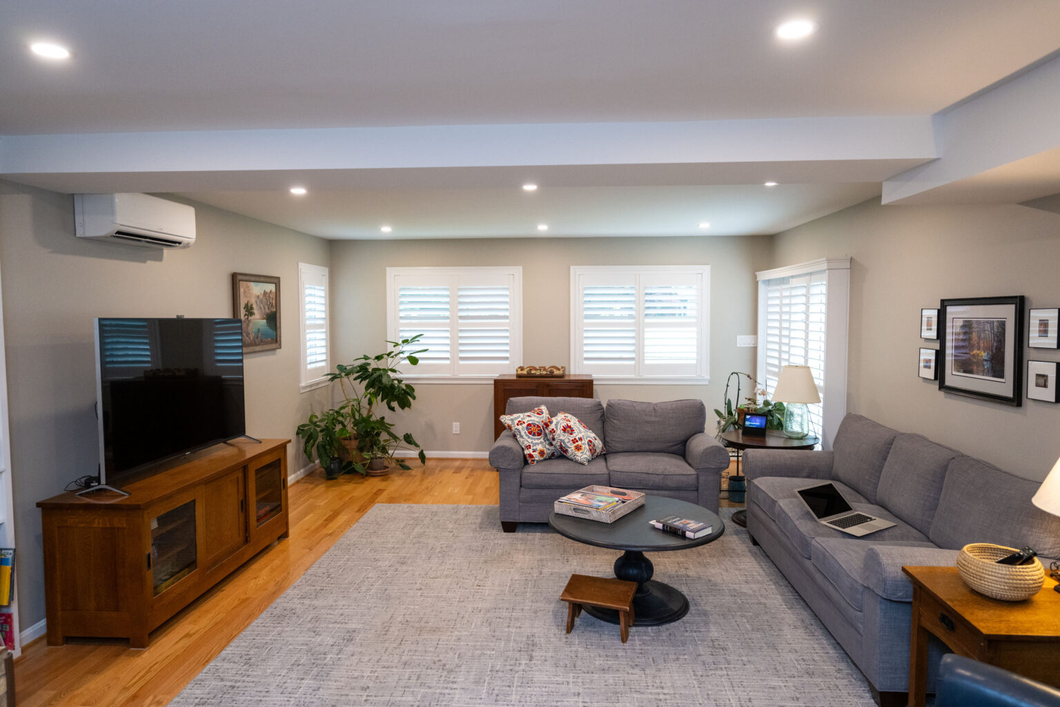 A living room with two gray sofas, a TV on a wooden stand, a round coffee table, a plant near the window, and framed pictures on the walls. The room has wooden floors and recessed lighting.