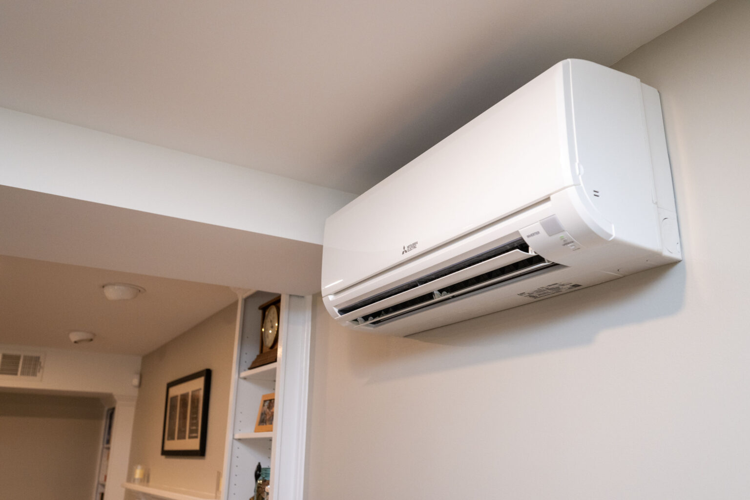 Wall-mounted white air conditioning unit installed indoors, blowing air into the room. Nearby corridor visible in the background with framed pictures on the walls.