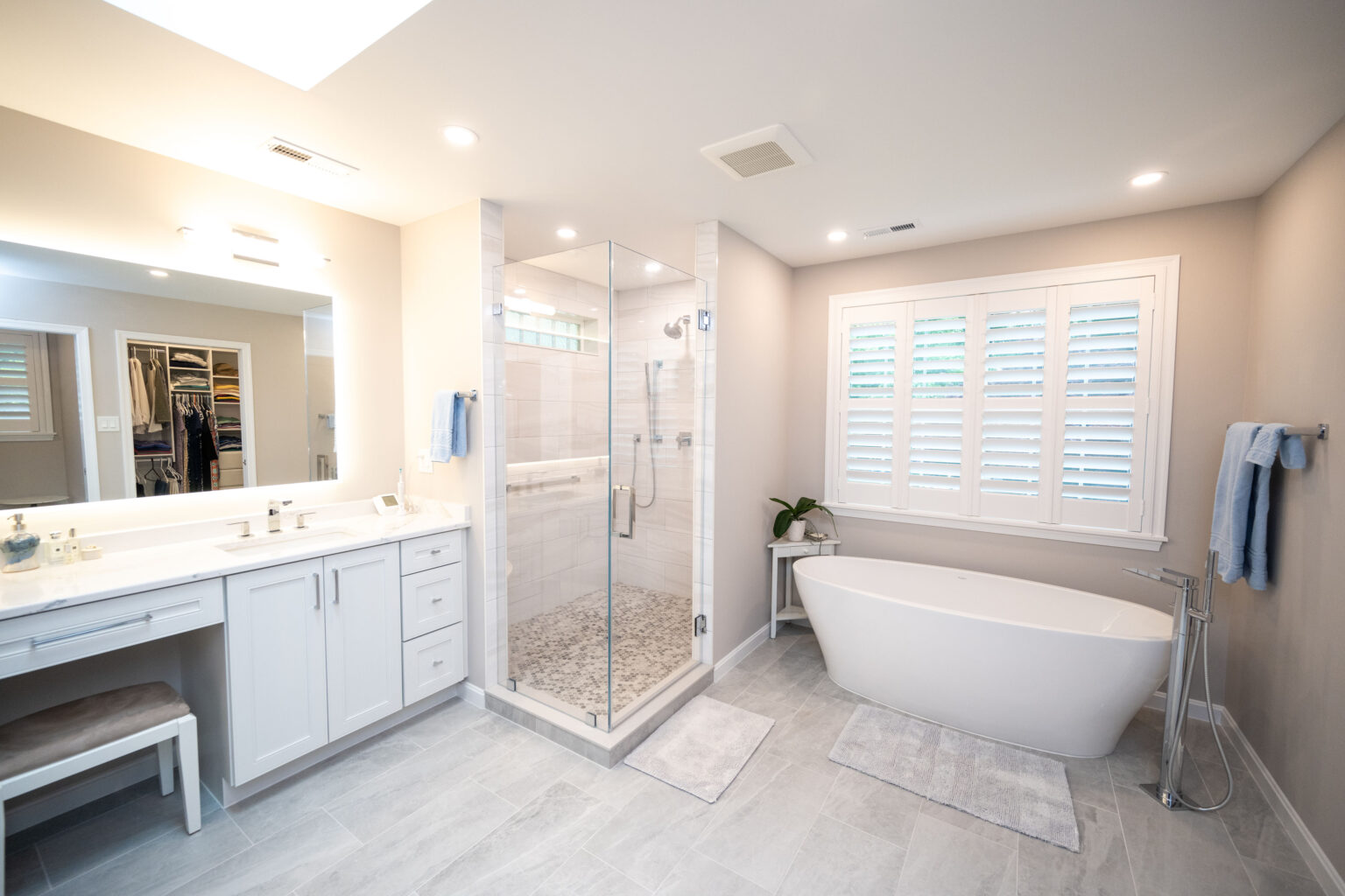 A modern bathroom with a glass-enclosed shower, freestanding bathtub, double sink vanity, and a window with white shutters. The space has beige walls, light grey tile flooring, and overhead lighting.