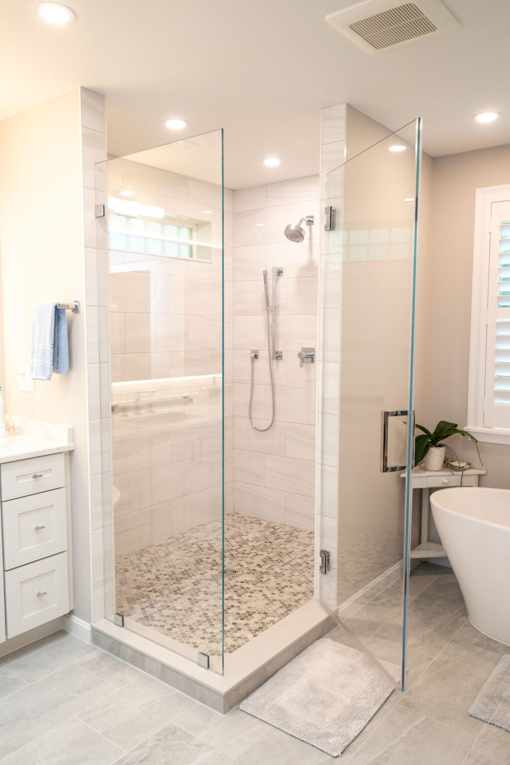 Modern bathroom with a glass-enclosed shower featuring a rainfall showerhead and handheld shower. The room includes tiled flooring, white cabinets, a freestanding bathtub, and a potted plant.