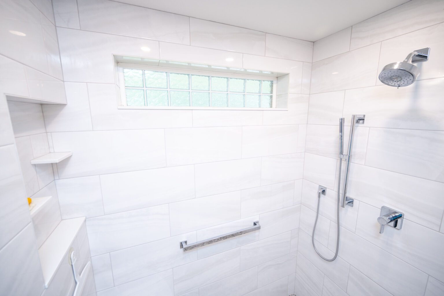 A modern bathroom shower with white tiled walls, featuring a glass block window, an overhead rainfall showerhead, and a handheld shower attachment.
