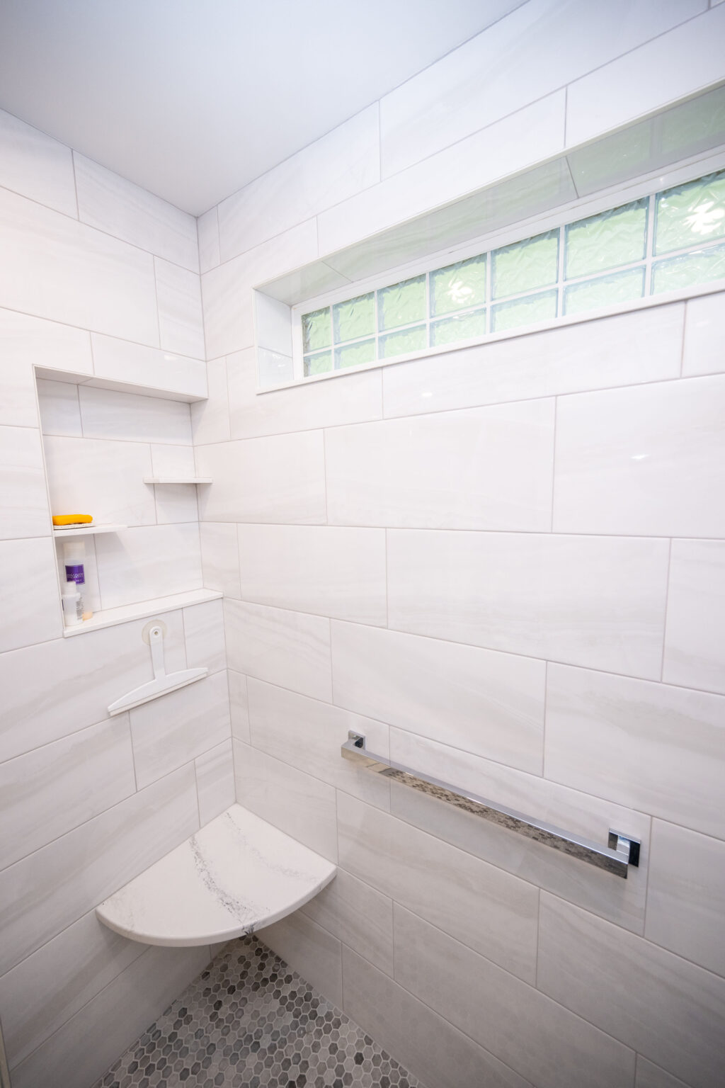 A shower with white tiled walls, a glass block window, corner shelves with toiletries, a small corner bench, and a towel bar. The floor has hexagonal mosaic tiles.