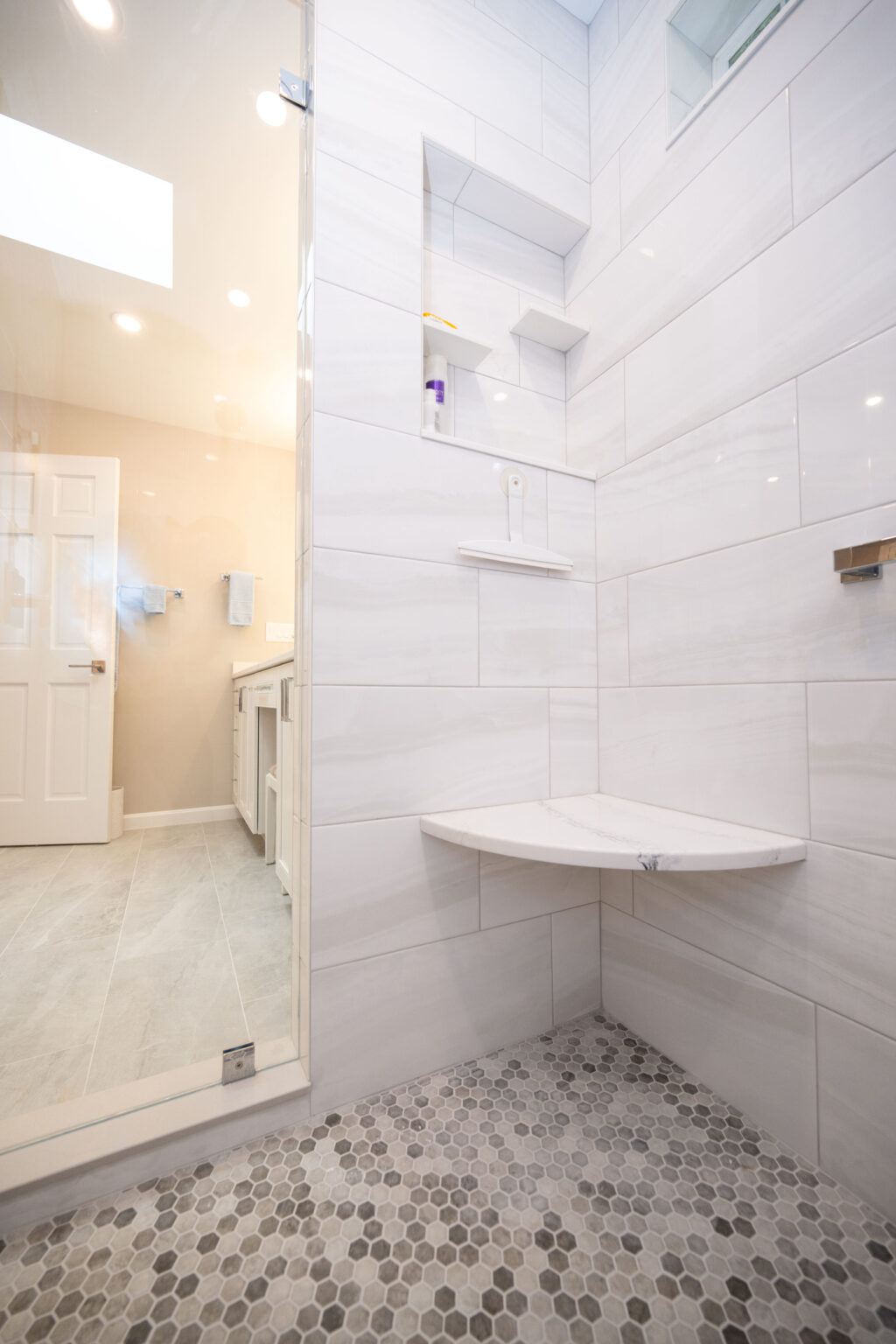 Modern bathroom with a glass-enclosed shower featuring white subway tiles, a corner bench, and built-in shelves. The bathroom also has a vanity with a mirror and a door in the background.