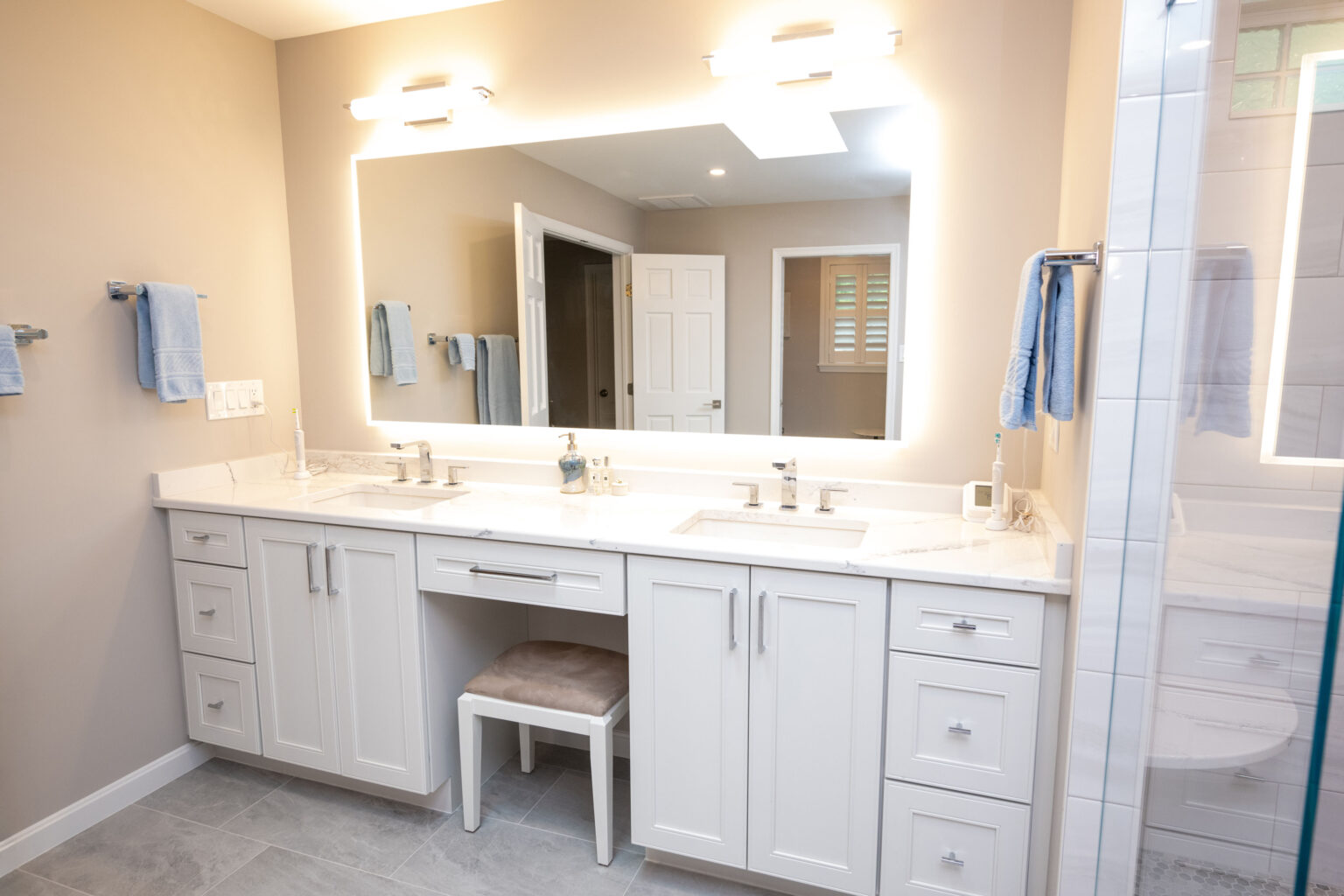 Modern bathroom with a large mirror, double sink vanity, white cabinets, a small stool underneath, and blue towels hung on the walls.