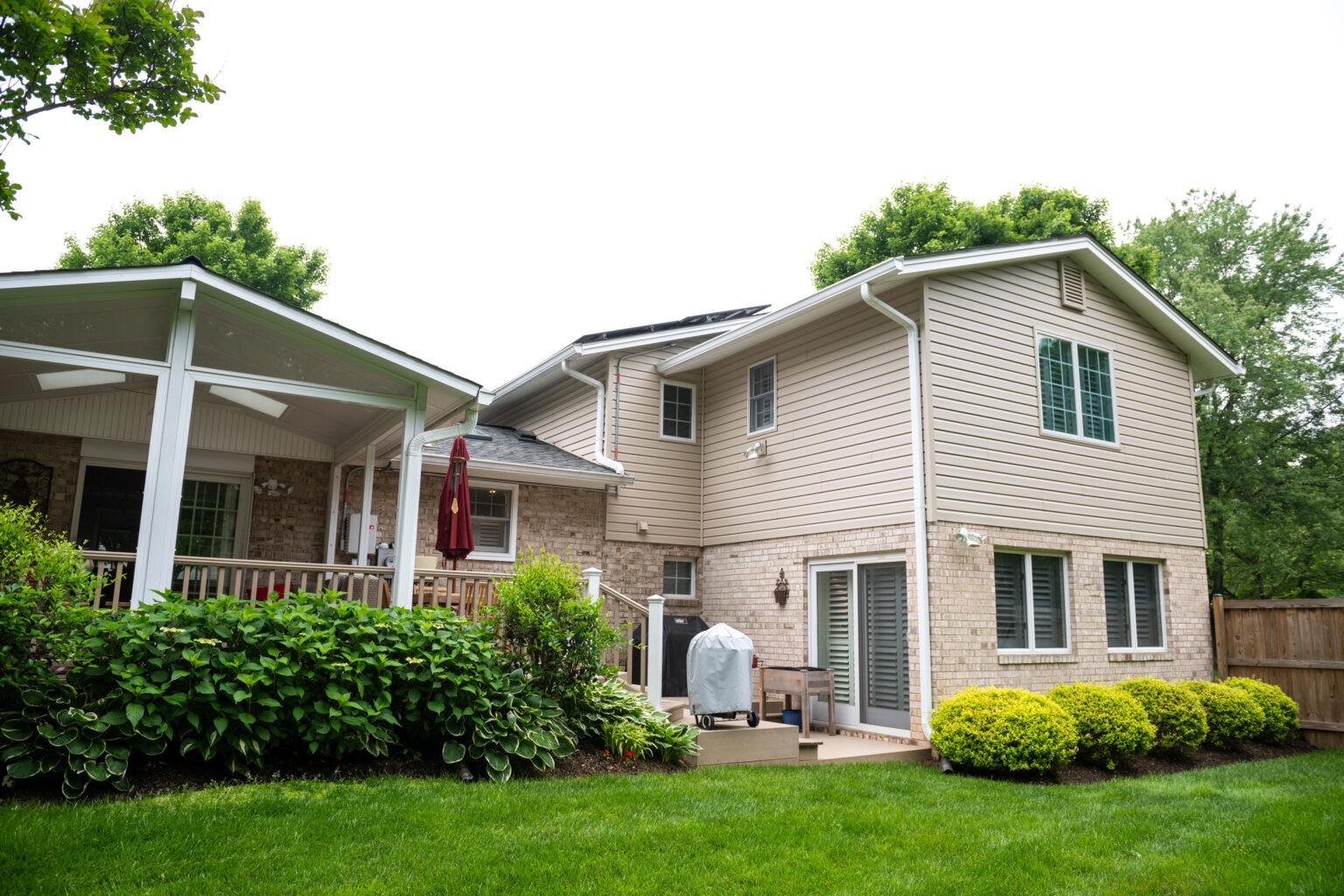 A backyard with a wooden deck, green lawn, shrubs, and various plants. A two-story house with beige siding and brick exterior is in the background. A grill and outdoor furniture are on the deck.