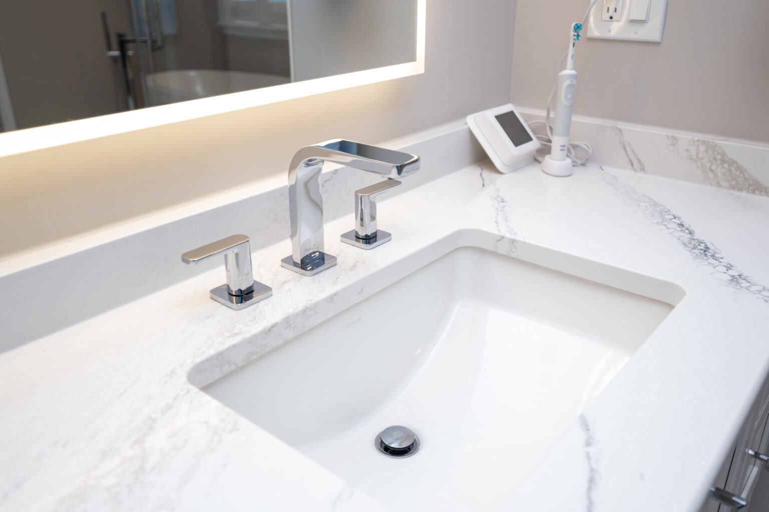 A modern bathroom sink with a rectangular basin, sleek chrome faucet, and handles on a white marble countertop. An electric toothbrush and a smart device are placed on the counter.