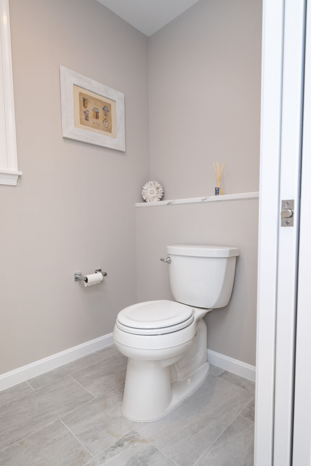 A small bathroom with a toilet, a roll of toilet paper on the holder, a decorative picture on the wall, and a small shelf with a diffuser and a white decorative item.