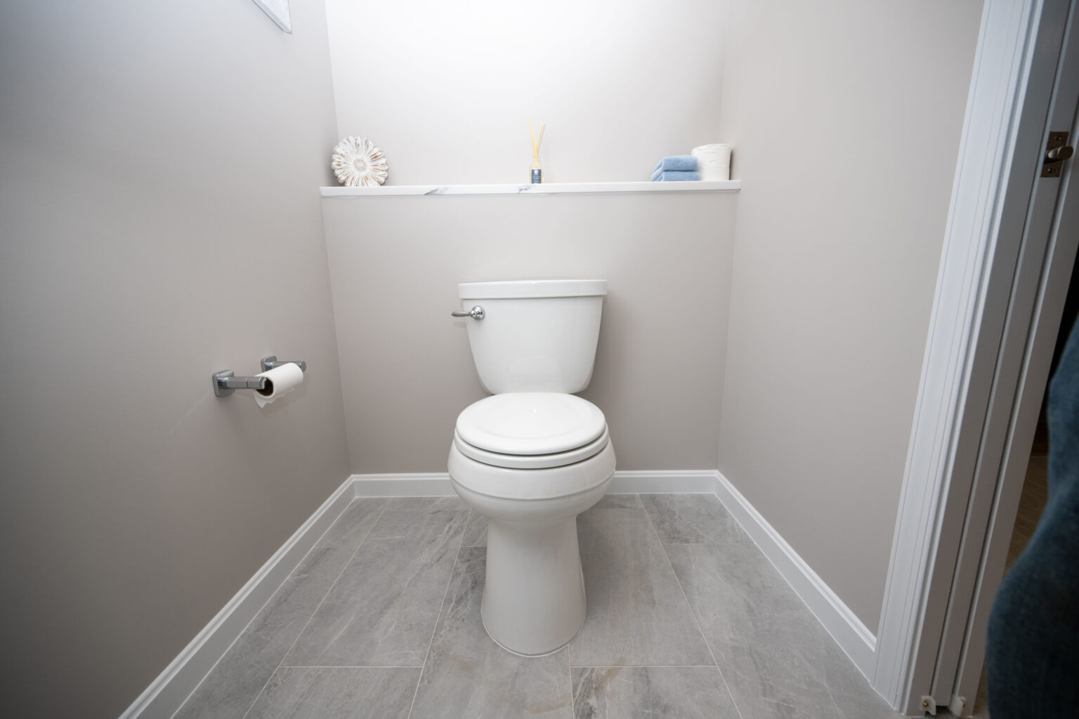A small, modern bathroom with a white toilet; a roll of toilet paper on the wall to the left, and decor items on a shelf behind the toilet. The floor is tiled.