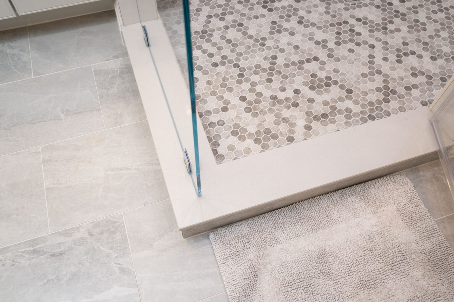 A bathroom with grey hexagonal tiles inside a glass shower enclosure, adjacent to light grey square floor tiles and a beige bath mat.
