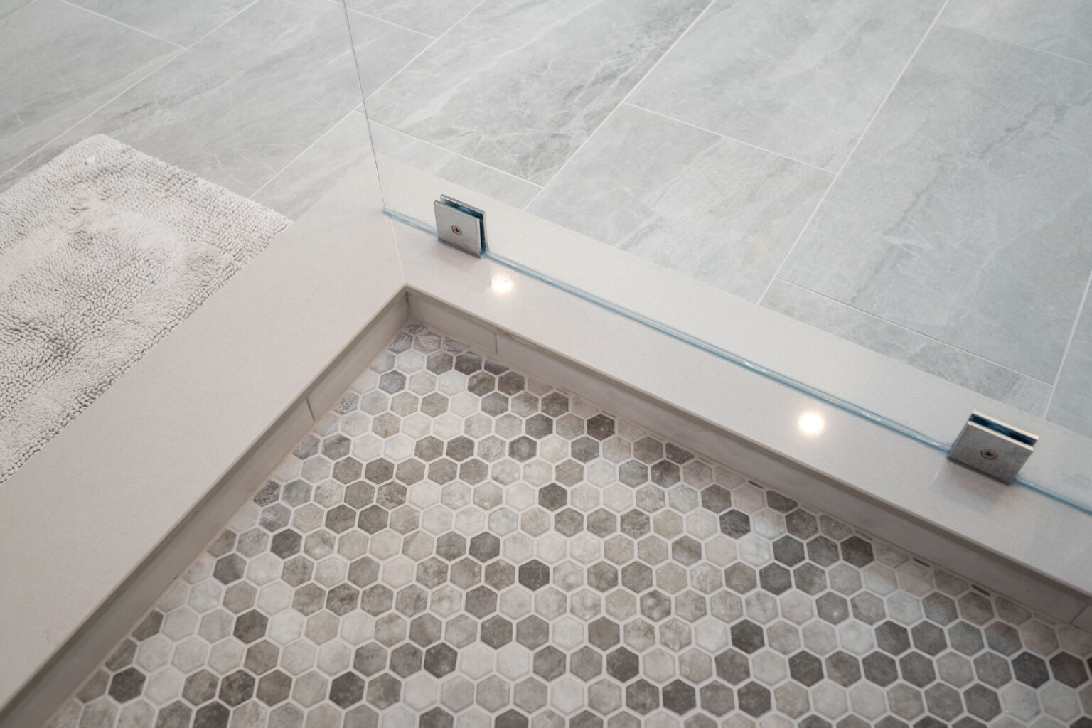 A shower area with hexagonal tile flooring, a frameless glass shower partition, and light-colored square floor tiles outside. A bath mat is partially visible.