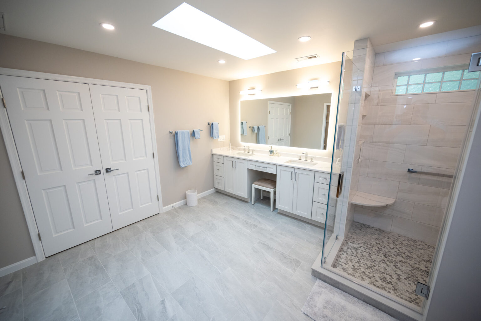 A modern bathroom features a glass-enclosed shower, a double-sink vanity with a large mirror, white cabinetry, towel racks with blue towels, and a double-door closet. The room has recessed lighting.