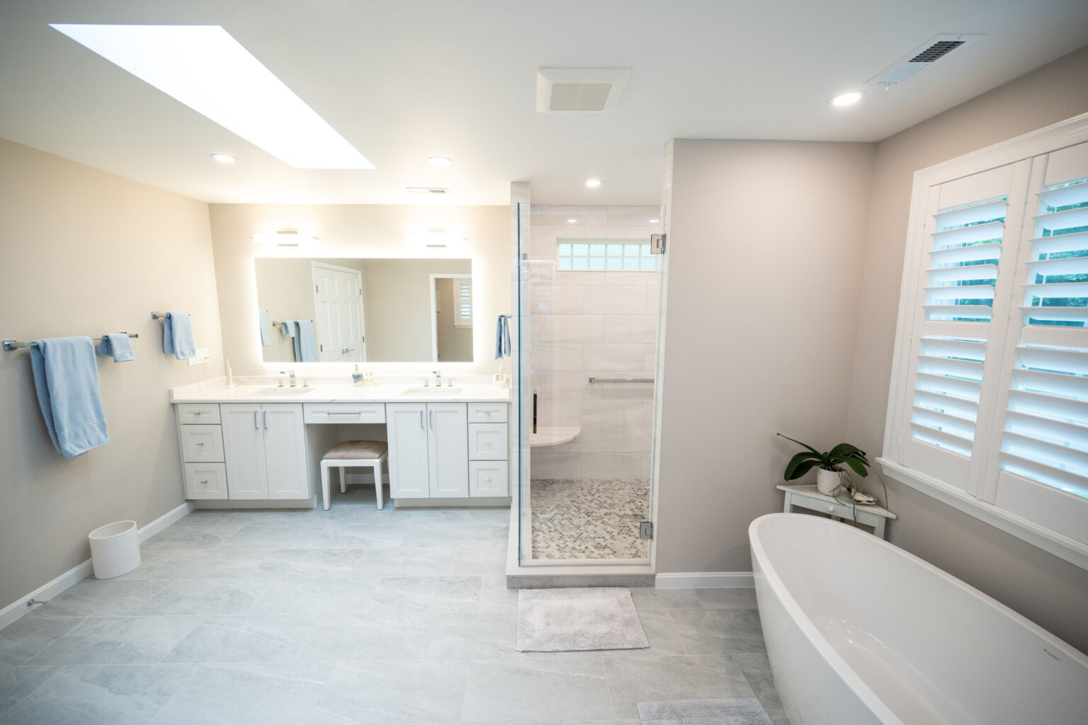 Modern bathroom with a double-sink vanity, large mirror, walk-in shower with glass door, freestanding bathtub, gray floor tiles, and white walls. The room is brightly lit with natural light.