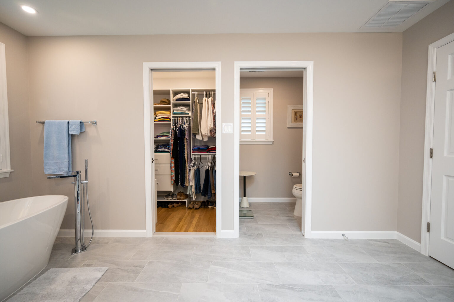 A modern bathroom features a bathtub on the left, a closet filled with clothes in the center, and a toilet in a separate room on the right. The floor is tiled in light grey.