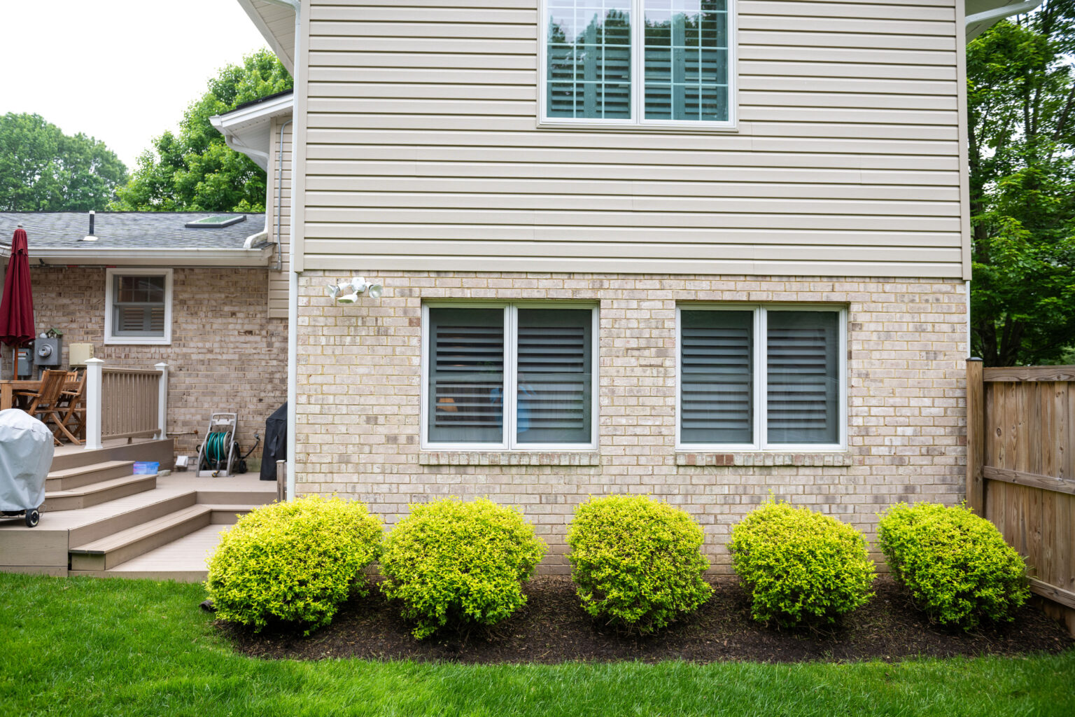 Backyard view of a beige brick house with two shuttered windows, a small deck with a grill and lawn tools, and four trimmed shrubs in front of a wooden fence.